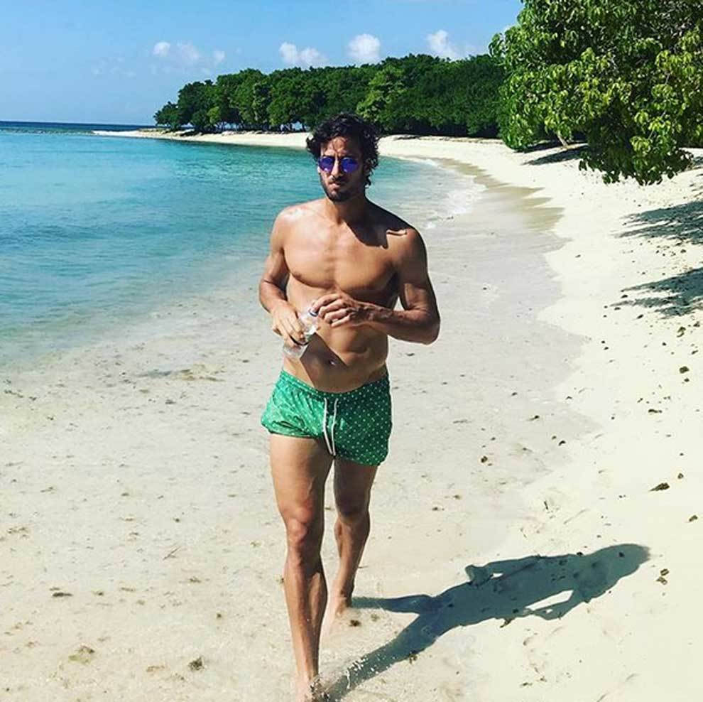 Man 桌布 壁紙 — 一個肌肉發達的男人穿著綠色泳褲和太陽眼鏡，赤腳走在白色沙灘上，背景是碧綠的海水和茂密的綠樹，天空湛藍。