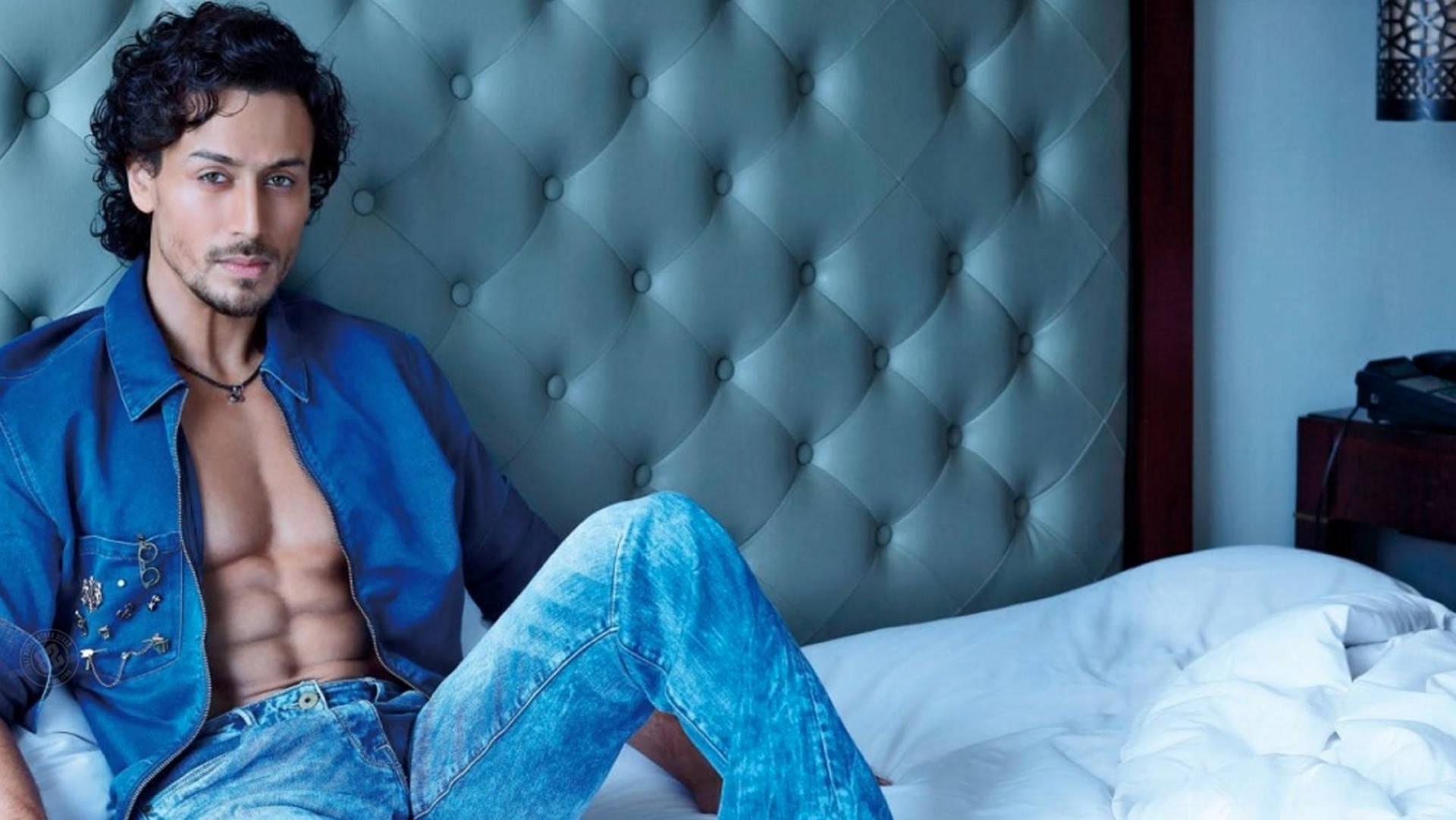 Tiger Shroff 桌布 壁紙 — 一個肌肉發達的男人穿著藍色牛仔褲，在冷藍燈光下，赤裸上身躺在一張有簇絨床頭板的床上。