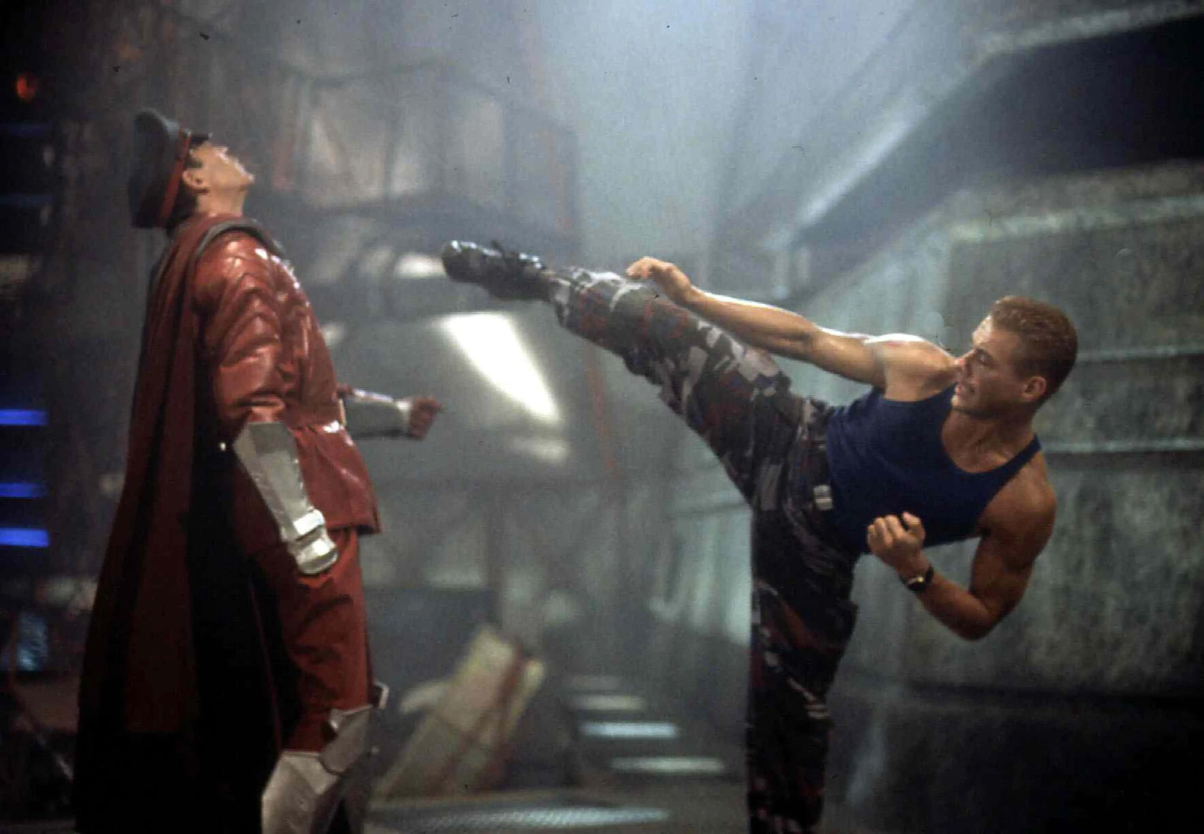 jean claude van damme 桌布 壁紙 — 一個肌肉發達的男人穿著迷彩褲，在煙霧瀰漫的工業環境中對一個穿著紅色外套和帽子的男人使出高踢腿。