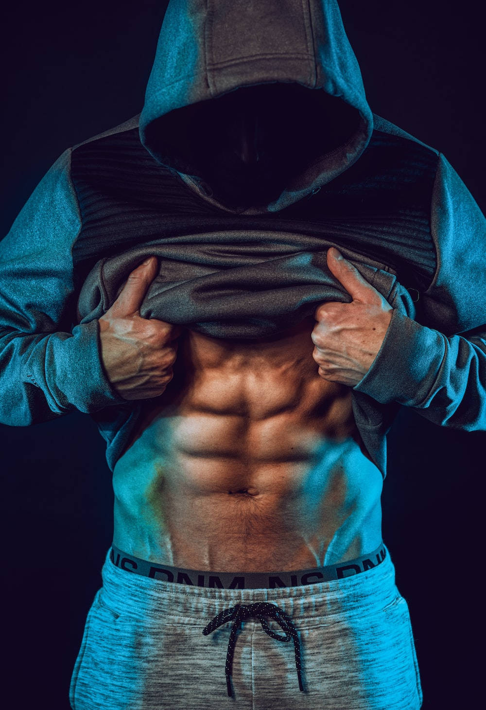 Exercise 桌布 壁紙 — 一個肌肉發達的男人穿著連帽衫，在戲劇性的藍色燈光下拉開衣服，露出線條分明的腹肌。