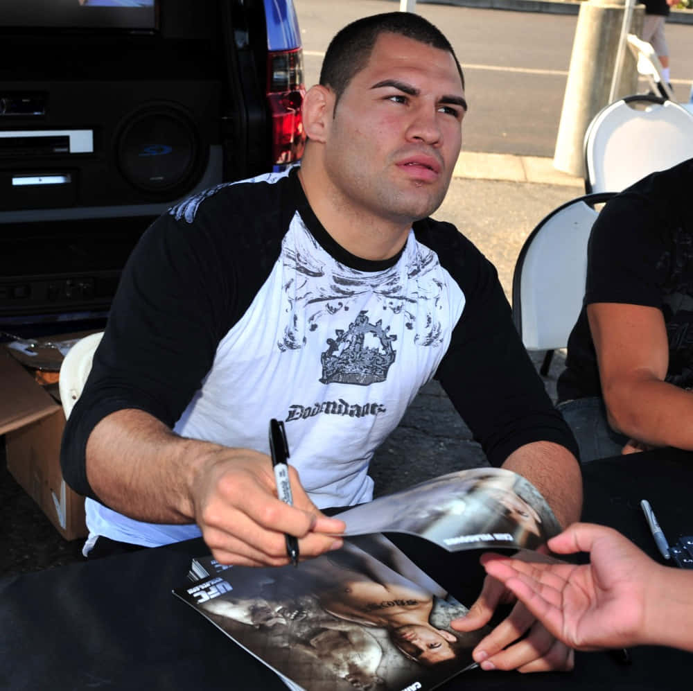 UFC 桌布 壁紙 — 一個肌肉發達的男人穿著黑白圖案T恤，在戶外一輛黑色SUV附近為粉絲簽署UFC雜誌。
