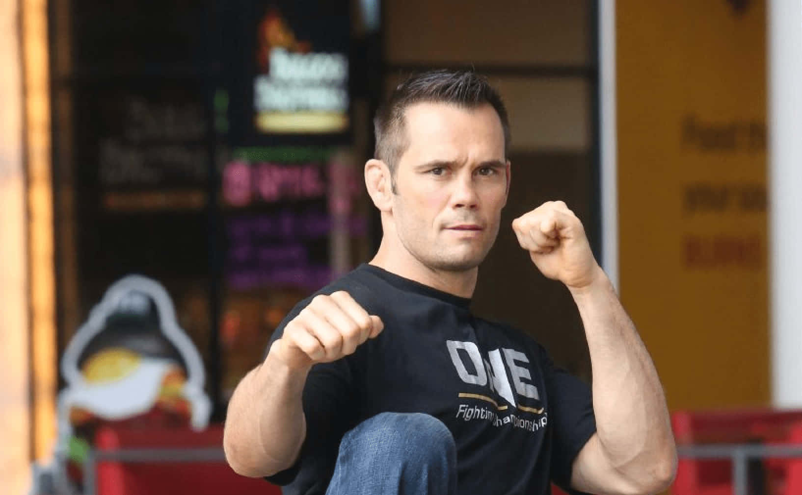 UFC 桌布 壁紙 — 一個肌肉發達的男人穿著黑色「ONE Fighting Championship」T恤，握緊拳頭以戰鬥姿勢坐在戶外。