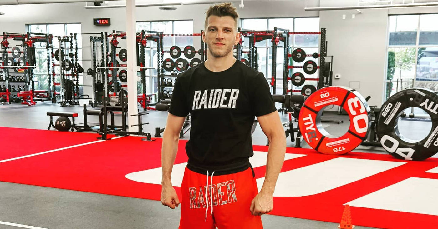 健身房 桌布 壁紙 — 一個肌肉發達的男人穿著黑色 'RAIDER' T恤和紅色短褲，自信地站在一個明亮的健身房裡，地板是紅色的，還有舉重設備。