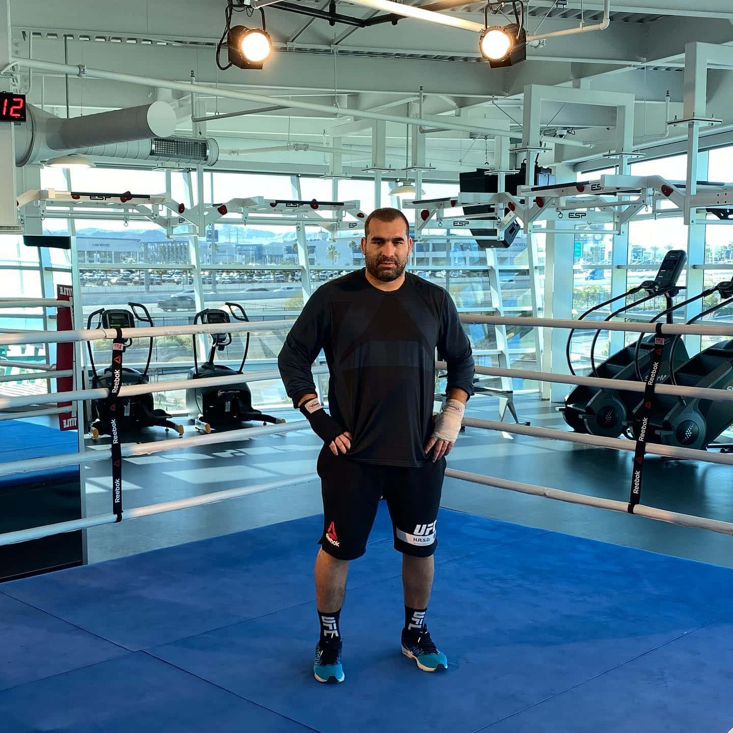 MMA 桌布 壁紙 — 一個肌肉發達的男人穿著黑色UFC裝備，自信地站在藍色拳擊擂台內，透過大窗戶可以看到城市景觀。
