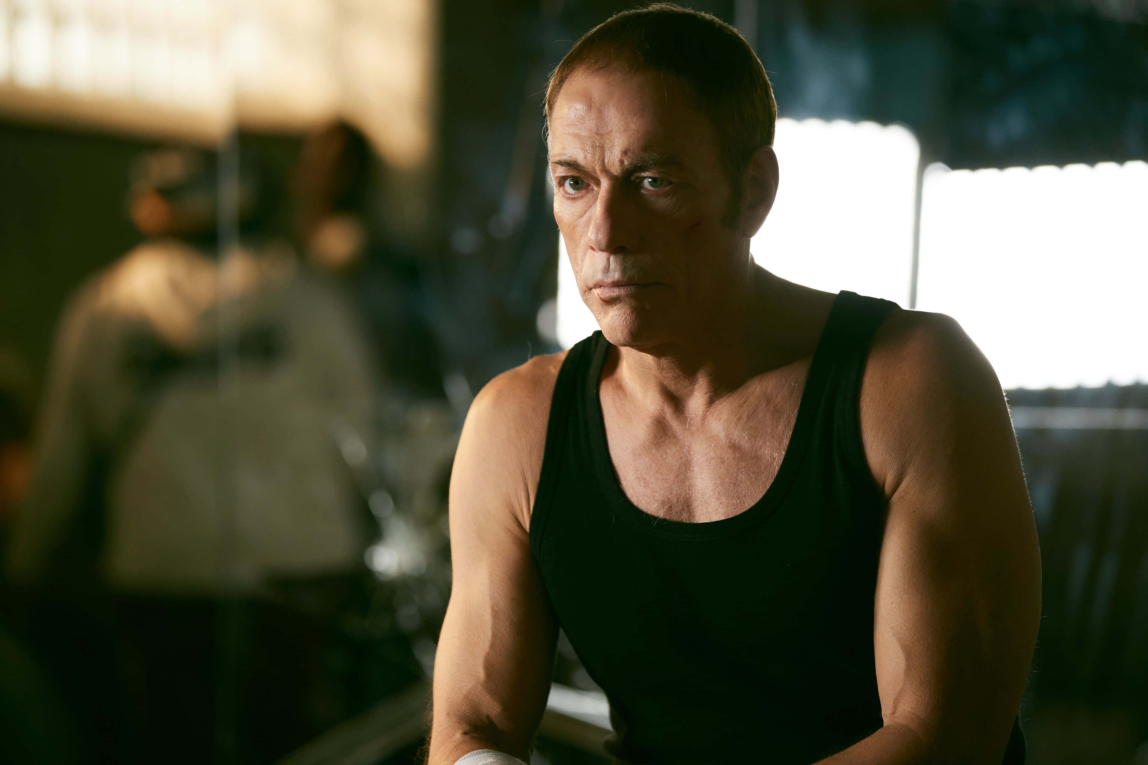 jean claude van damme 桌布 壁紙 — 一個肌肉發達的男人穿著黑色背心，在光線昏暗的健身房裡，陽光透過窗戶照射進來，他凝視著前方。