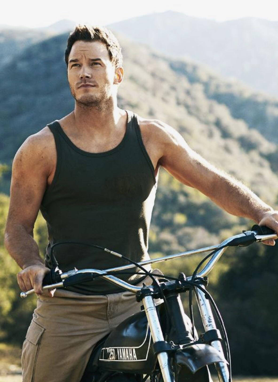 Chris Pratt 桌布 壁紙 — 一個肌肉發達的男人穿著黑色背心，靠在一輛山葉摩托車上，背景是陽光明媚的山脈。