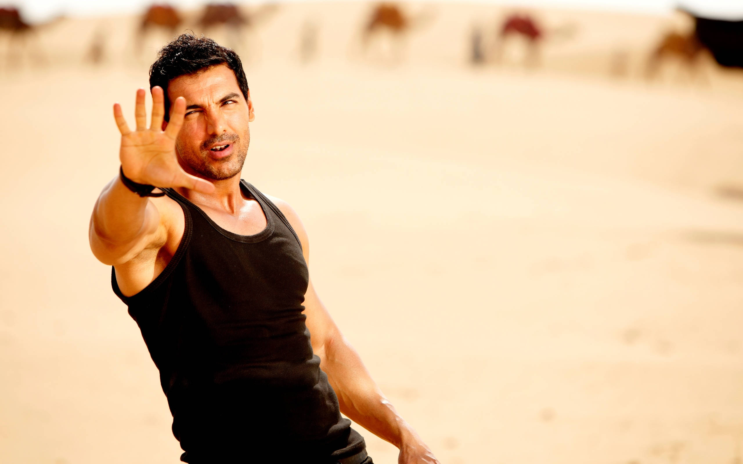 John Abraham 桌布 壁紙 — 一個肌肉發達的男人穿著黑色背心，在背景有駱駝的沙漠中向前揮手。