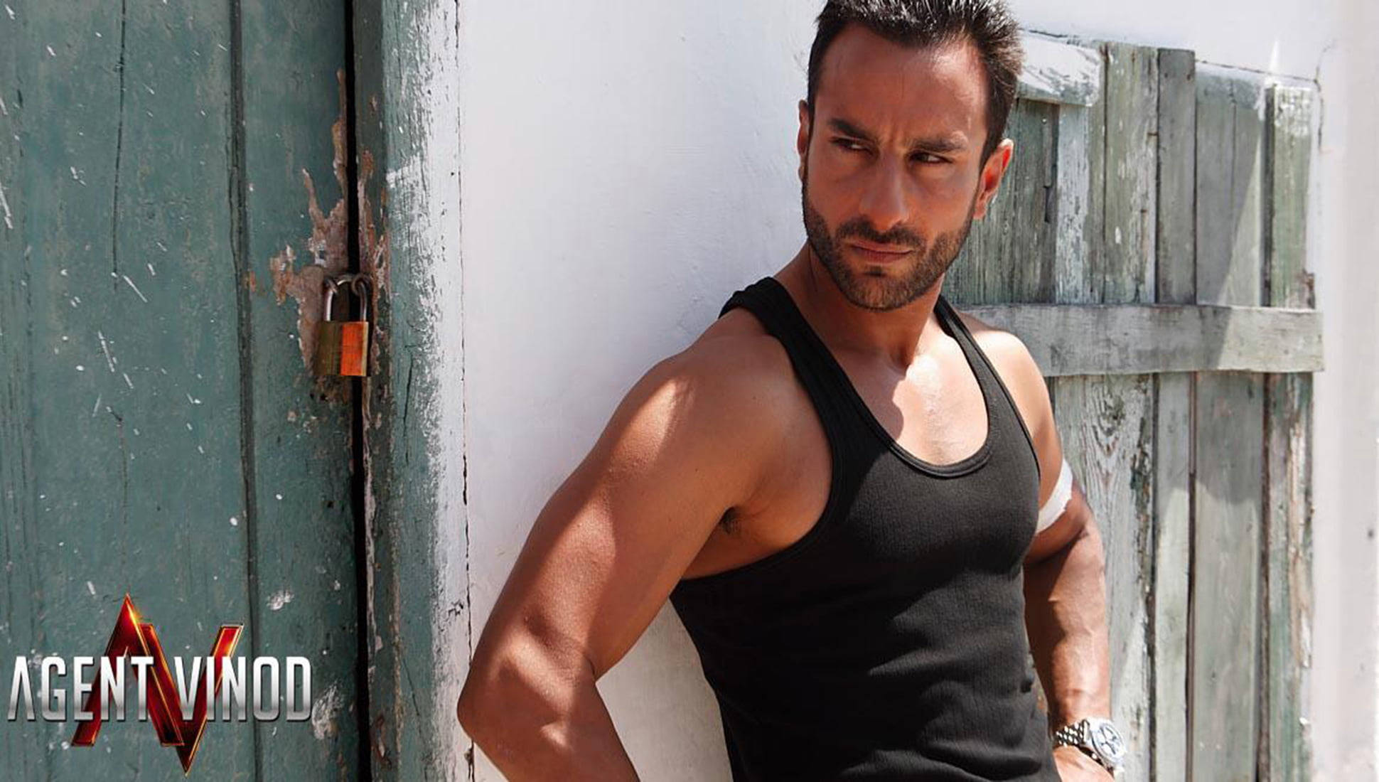 Saif Ali Khan 桌布 壁紙 — 一個肌肉發達的男人穿著黑色背心，在明亮的陽光下靠在一扇帶有生鏽掛鎖的風化綠色門上。