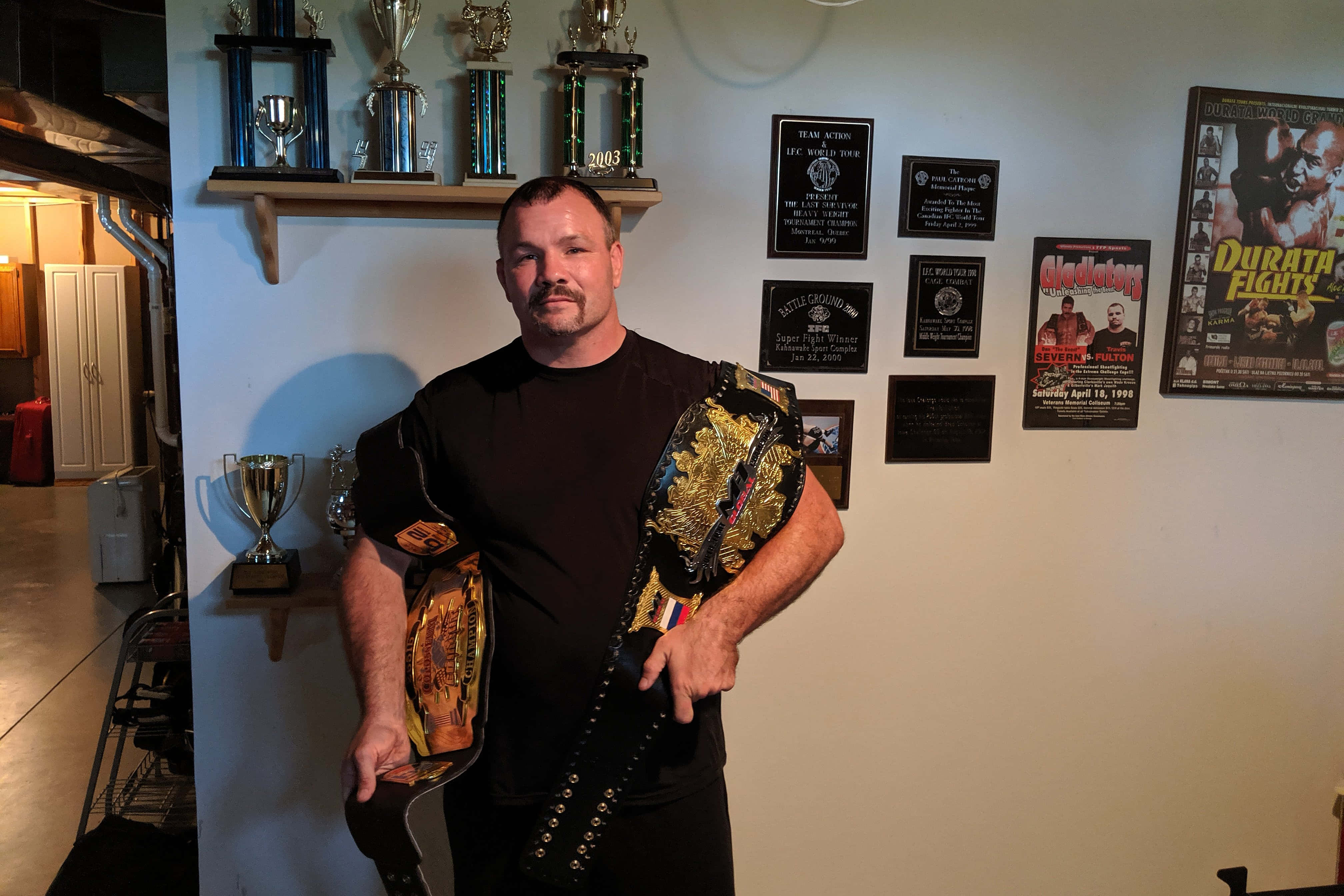 UFC 桌布 壁紙 — 一個肌肉發達的男人穿著黑色襯衫，在一個充滿獎盃的車庫裡自豪地舉著冠軍腰帶，牆上掛著裱框的搏擊海報。