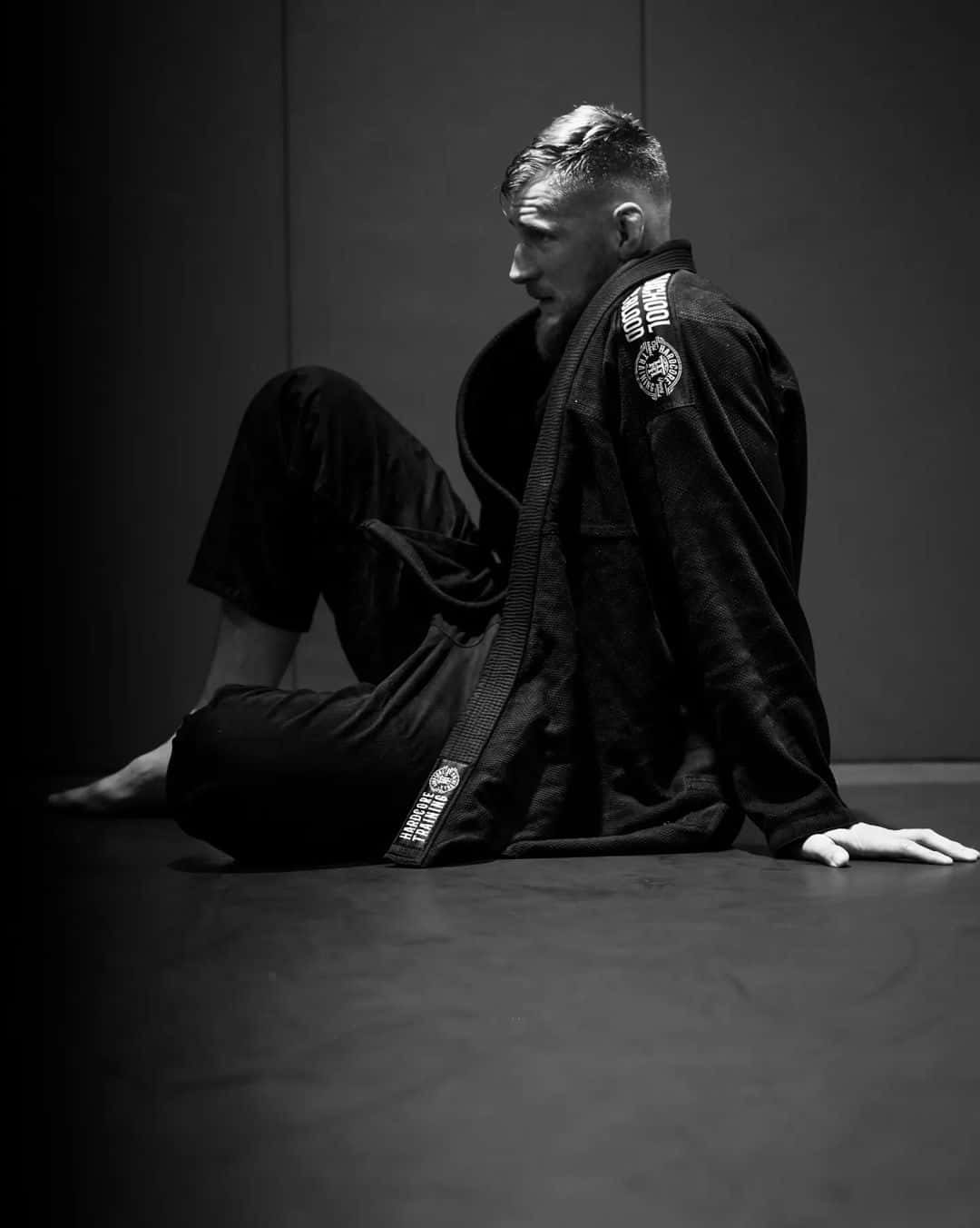 Athletic 桌布 壁紙 — 一個肌肉發達的男人穿著黑色道服，坐在光線昏暗的練習室的墊子上，目光專注地望向鏡頭外。
