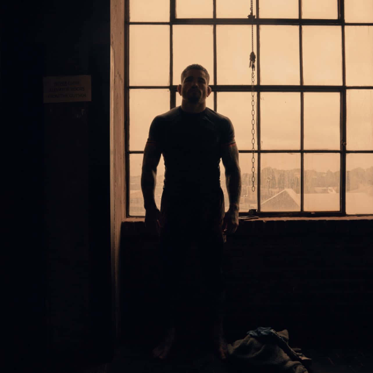 Silhouette 桌布 壁紙 — 一個肌肉發達的男人站在一個大型工業窗戶前形成剪影，金色的光線灑滿灰塵飛揚的房間。