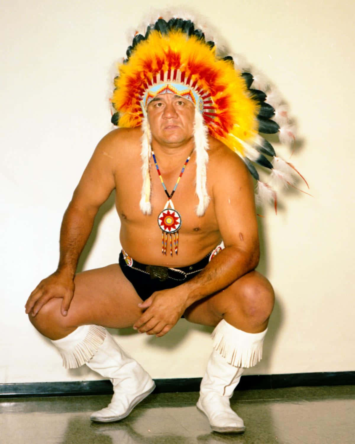 WWE 桌布 壁紙 — 一個肌肉發達的男子，戴著鮮豔的羽毛頭飾，穿著白色流蘇靴，蹲在素牆前。
