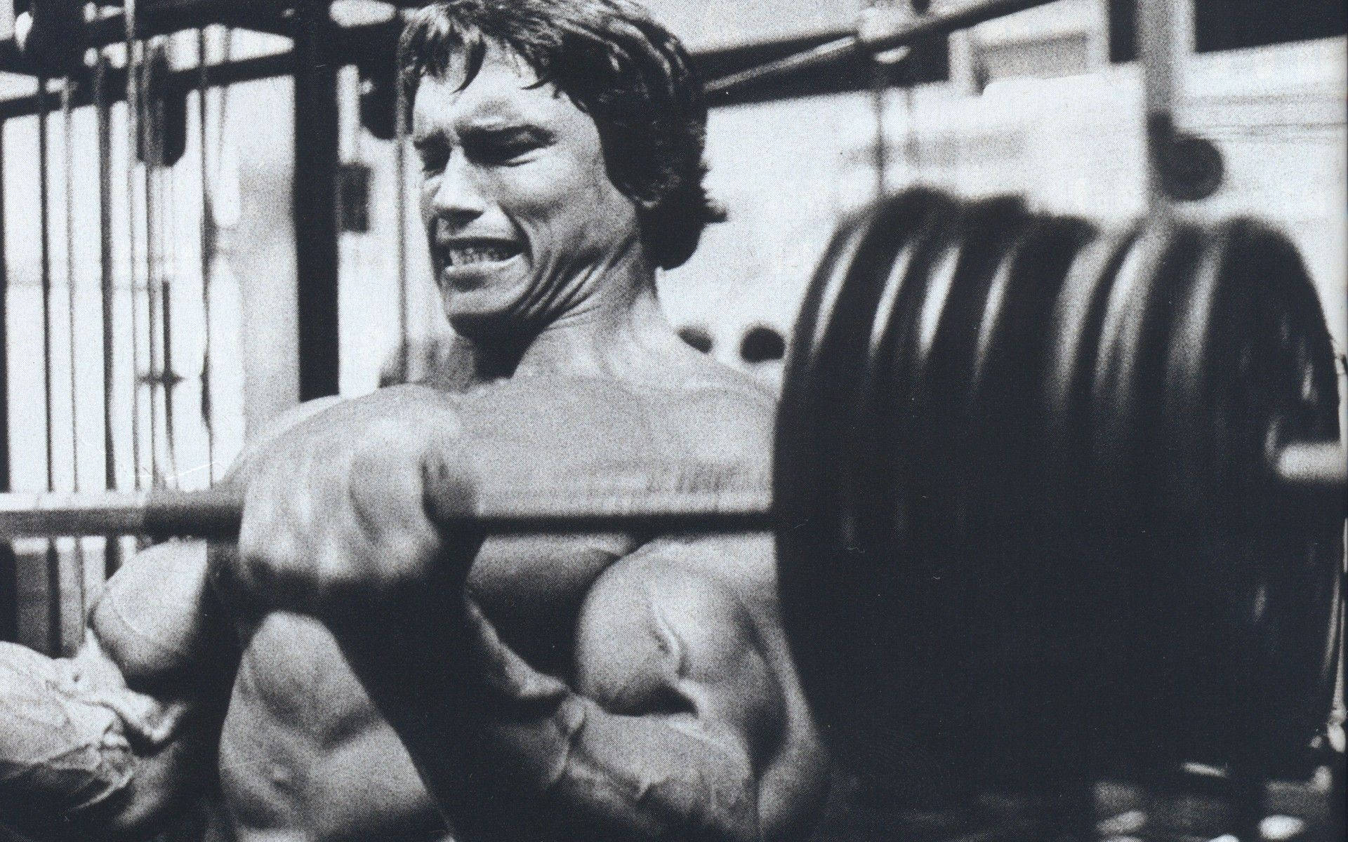 Arnold 桌布 壁紙 — 一個肌肉發達的男子在健身房用力舉起沉重的槓鈴，以粗糙的黑白捕捉。
