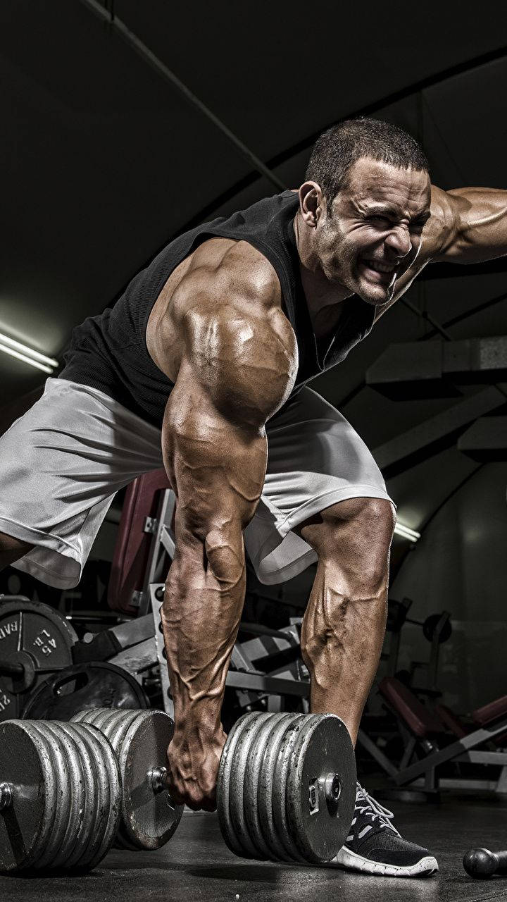 Exercise 桌布 壁紙 — 一個肌肉發達的男子在光線昏暗的健身房內用力舉起沉重的槓鈴，周圍可見各種器材。