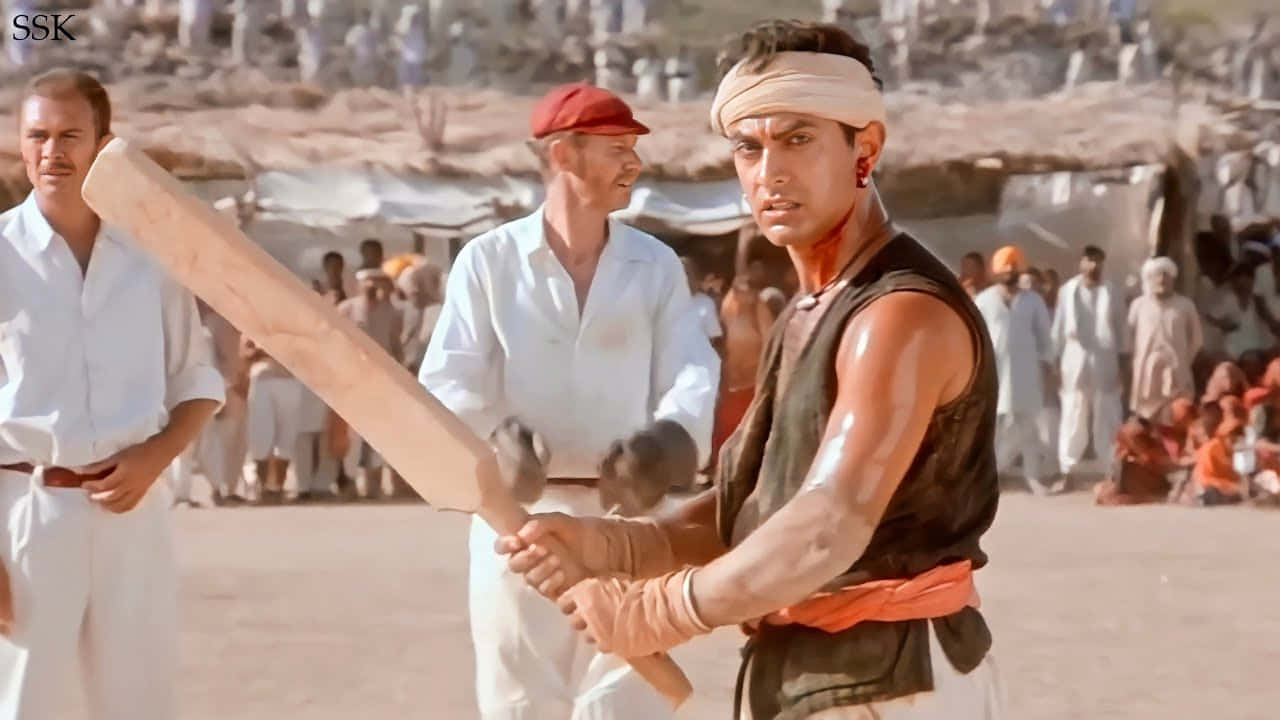 Aamir Khan 桌布 壁紙 — 一個肌肉發達的男子在塵土飛揚的場地上用力握著板球棒，觀眾在烈日下觀看。