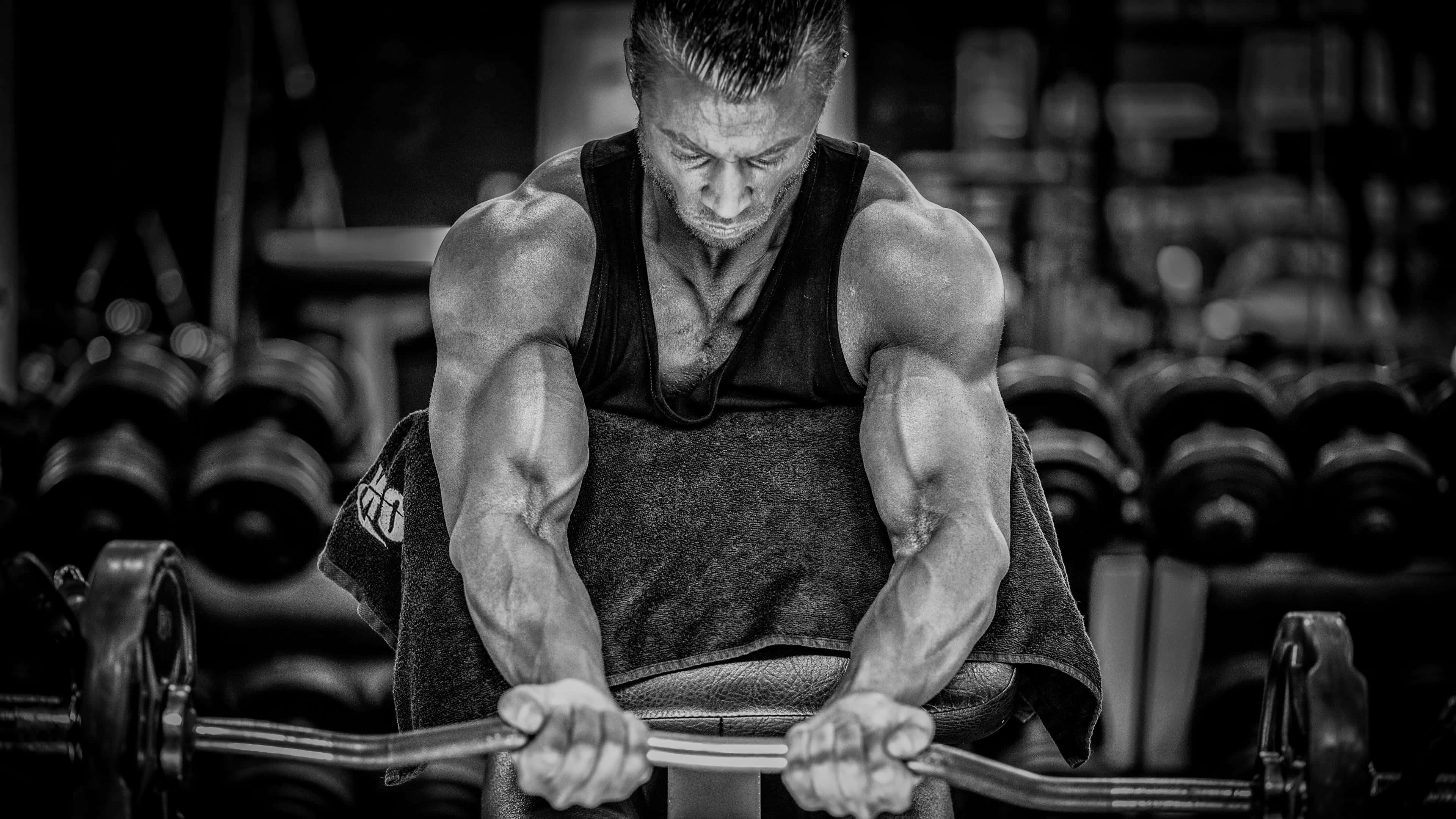 bodybuilding 桌布 壁紙 — 一個肌肉發達的男子在昏暗的健身房中用力舉起槓鈴，背景有模糊的啞鈴。