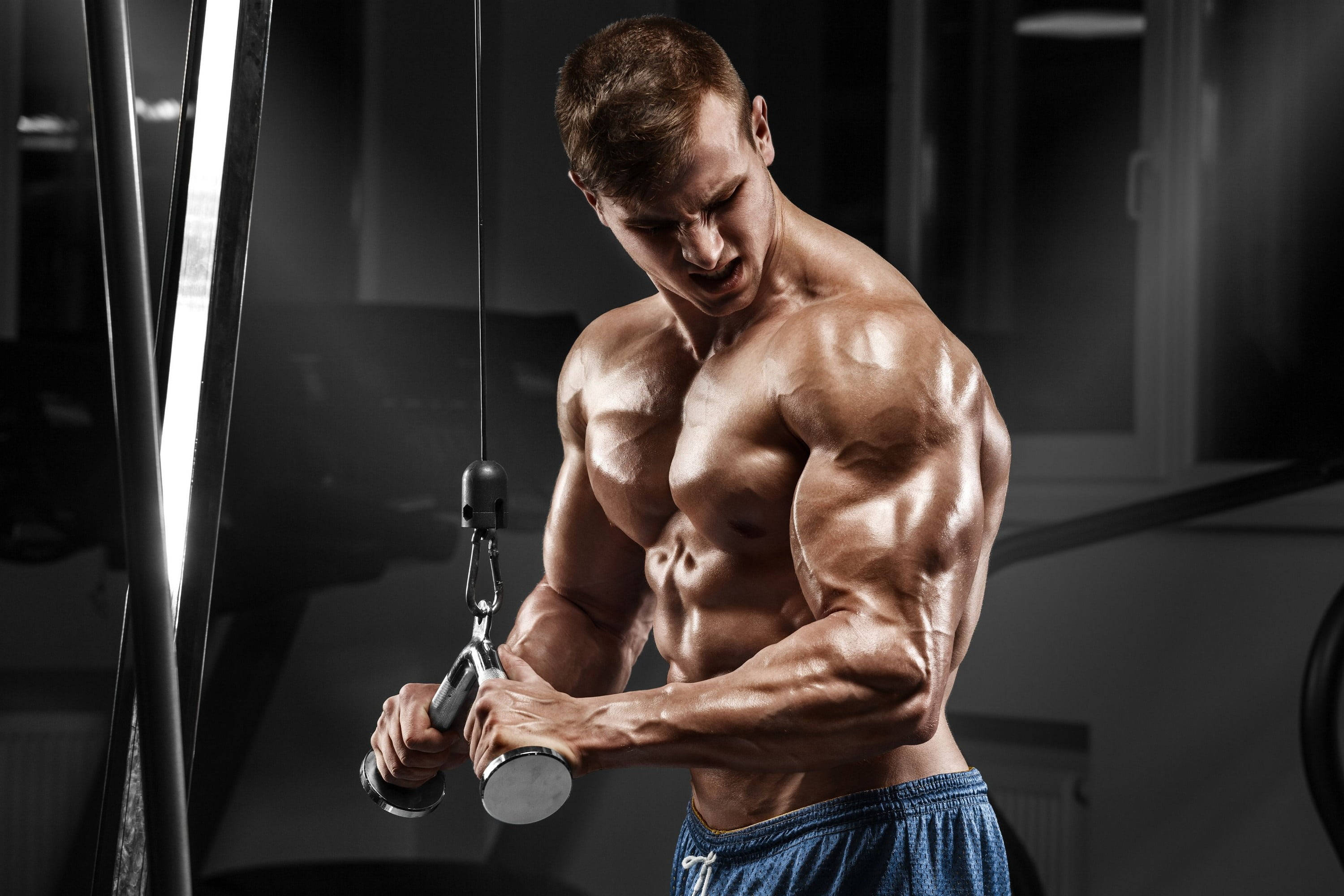 muscle growth 桌布 壁紙 — 一個肌肉發達的男子在燈光昏暗的健身房中，在戲劇性的燈光下用力拉起纜繩重量。
