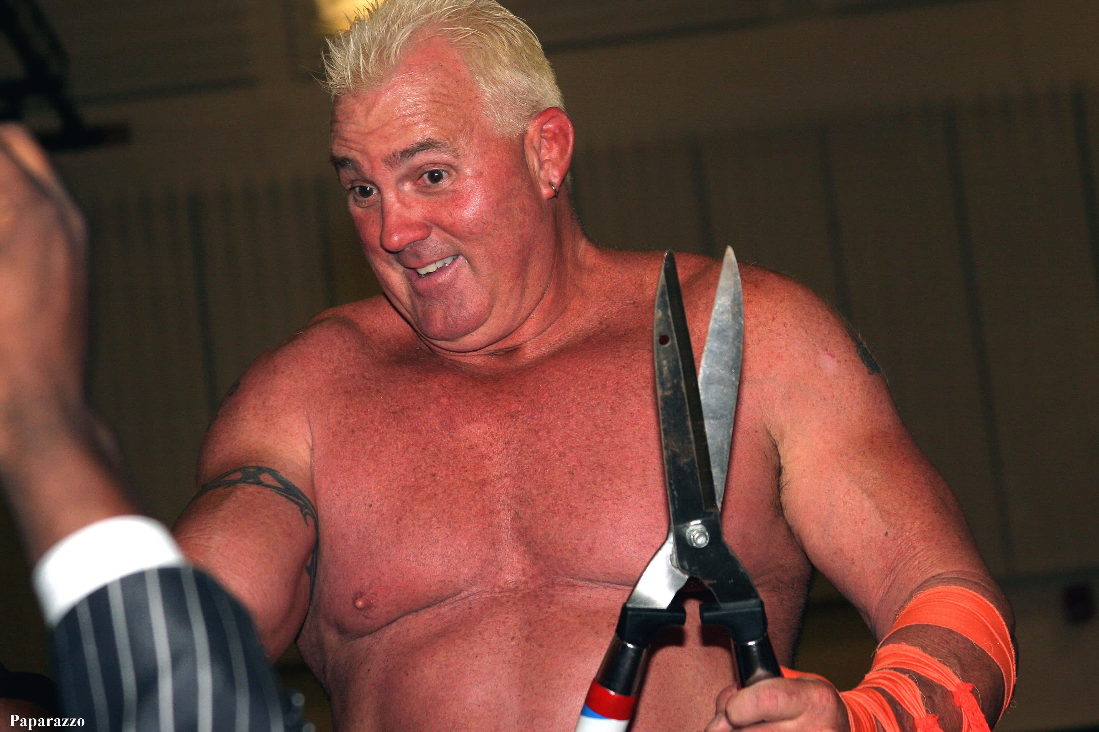 WWE 桌布 壁紙 — 一個肌肉發達的金髮男人，戴著橙色護腕，拿著一把巨大的剪刀，對著一個穿著細條紋西裝的人微笑。
