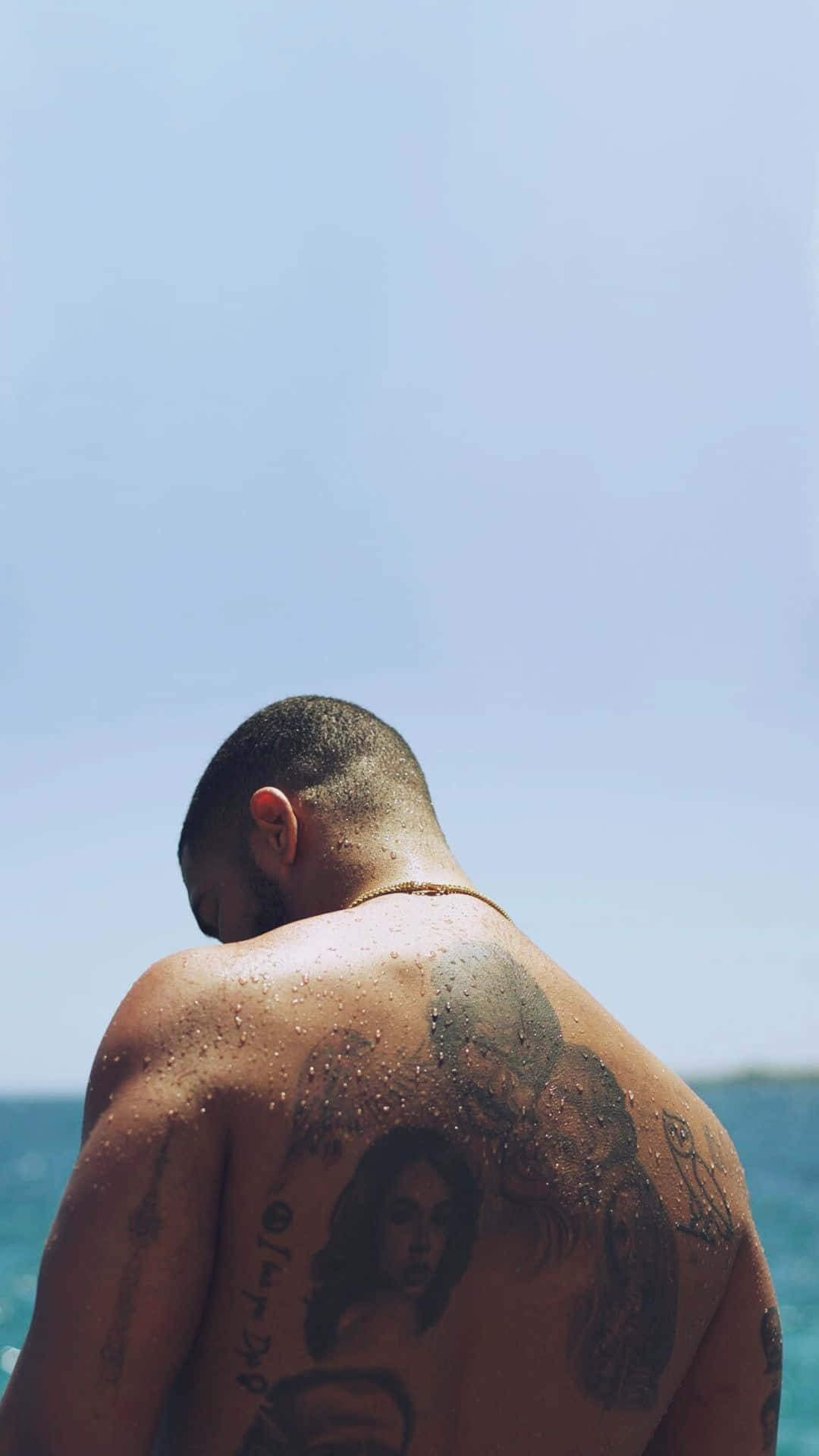 Aesthetic Drake 桌布 壁紙 — 一個背上有刺青的男人，在清澈的藍天下赤膊站在海邊，皮膚上有水滴。
