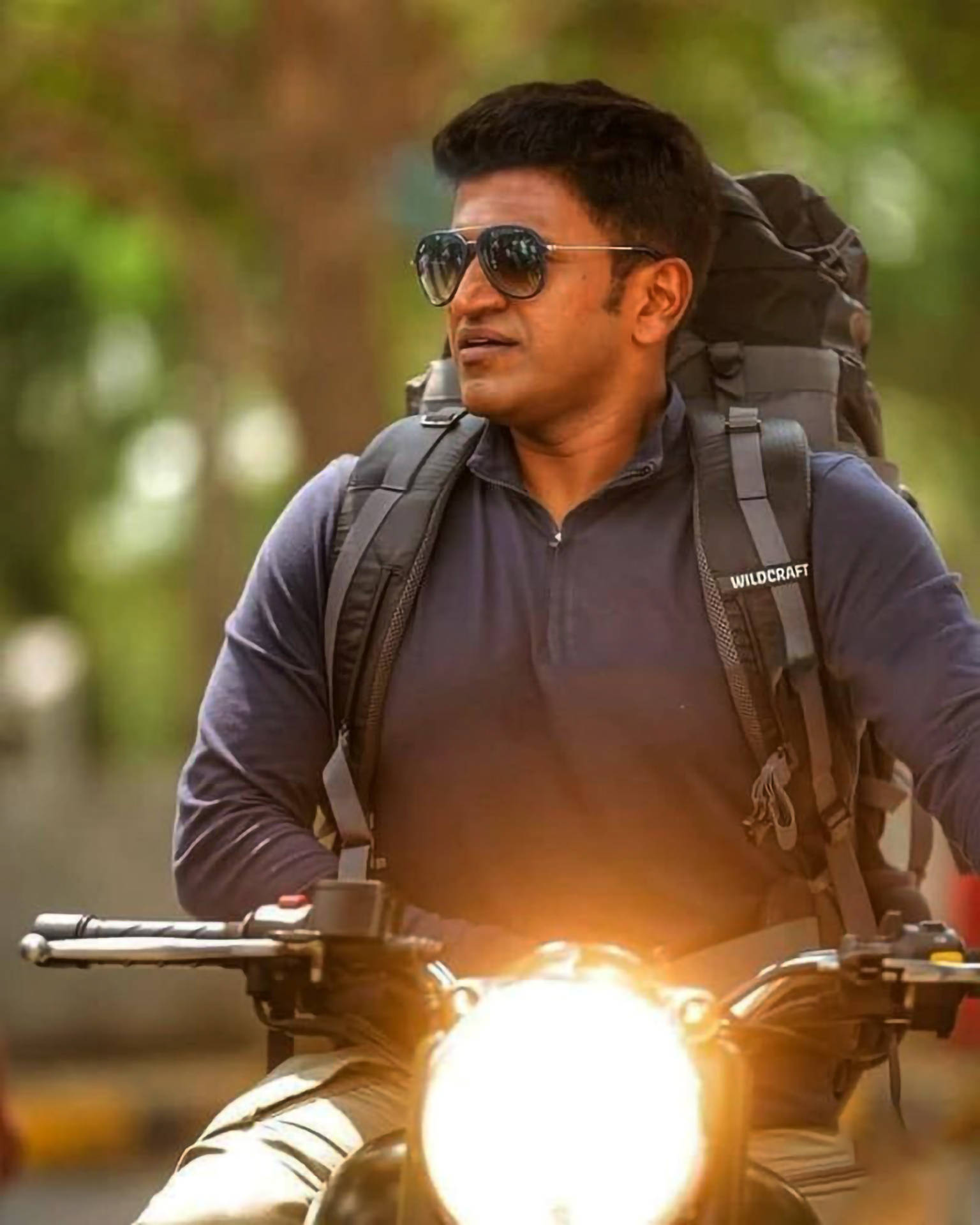 Puneeth Rajkumar 桌布 壁紙 — 一個背著大背包的男人騎著摩托車，明亮的車頭燈在模糊的綠色背景中發光。