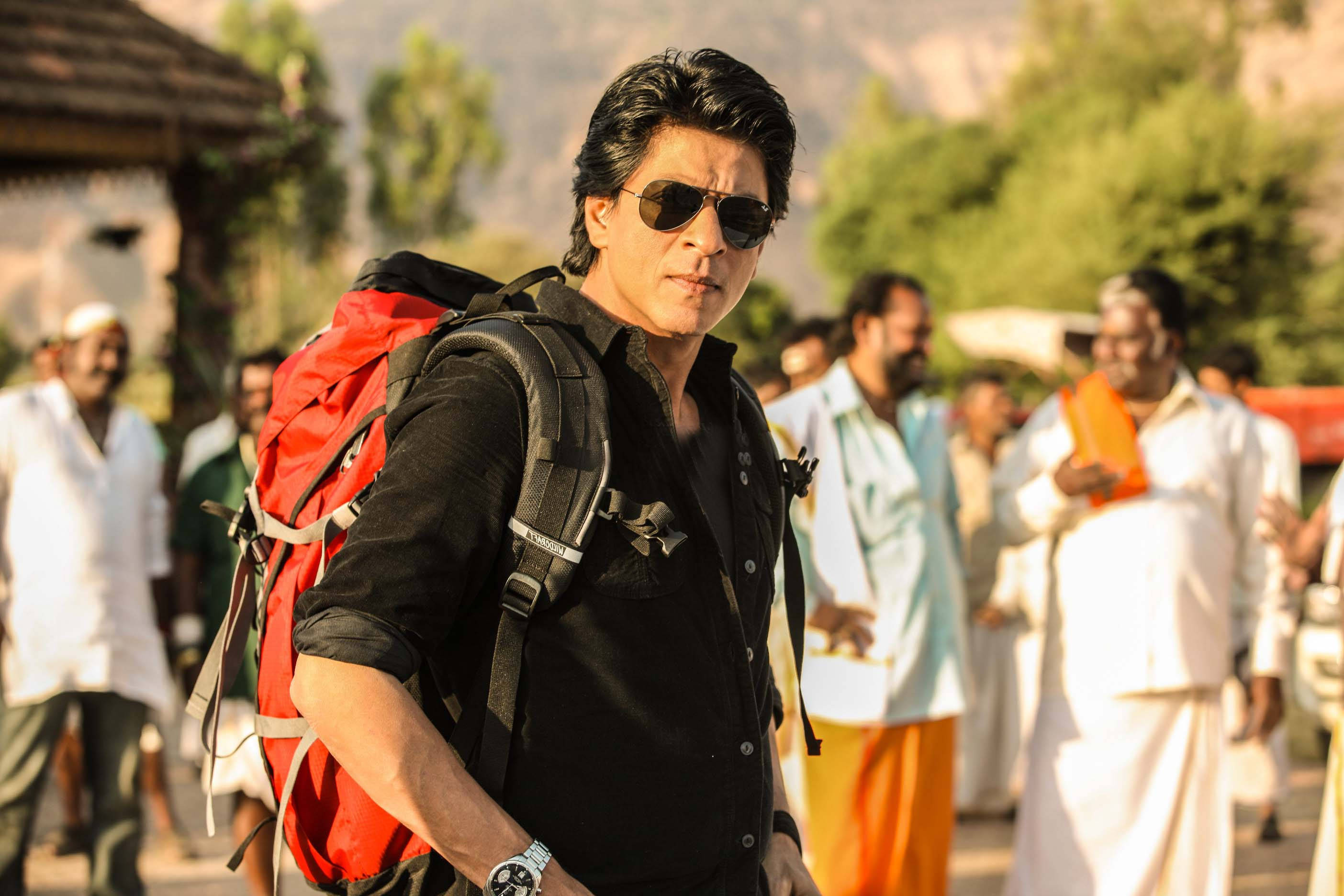 Shahrukh 桌布 壁紙 — 一個背著紅色背包、戴著太陽眼鏡的男人自信地走過一條陽光明媚的村莊街道，背景中有模糊的當地人。
