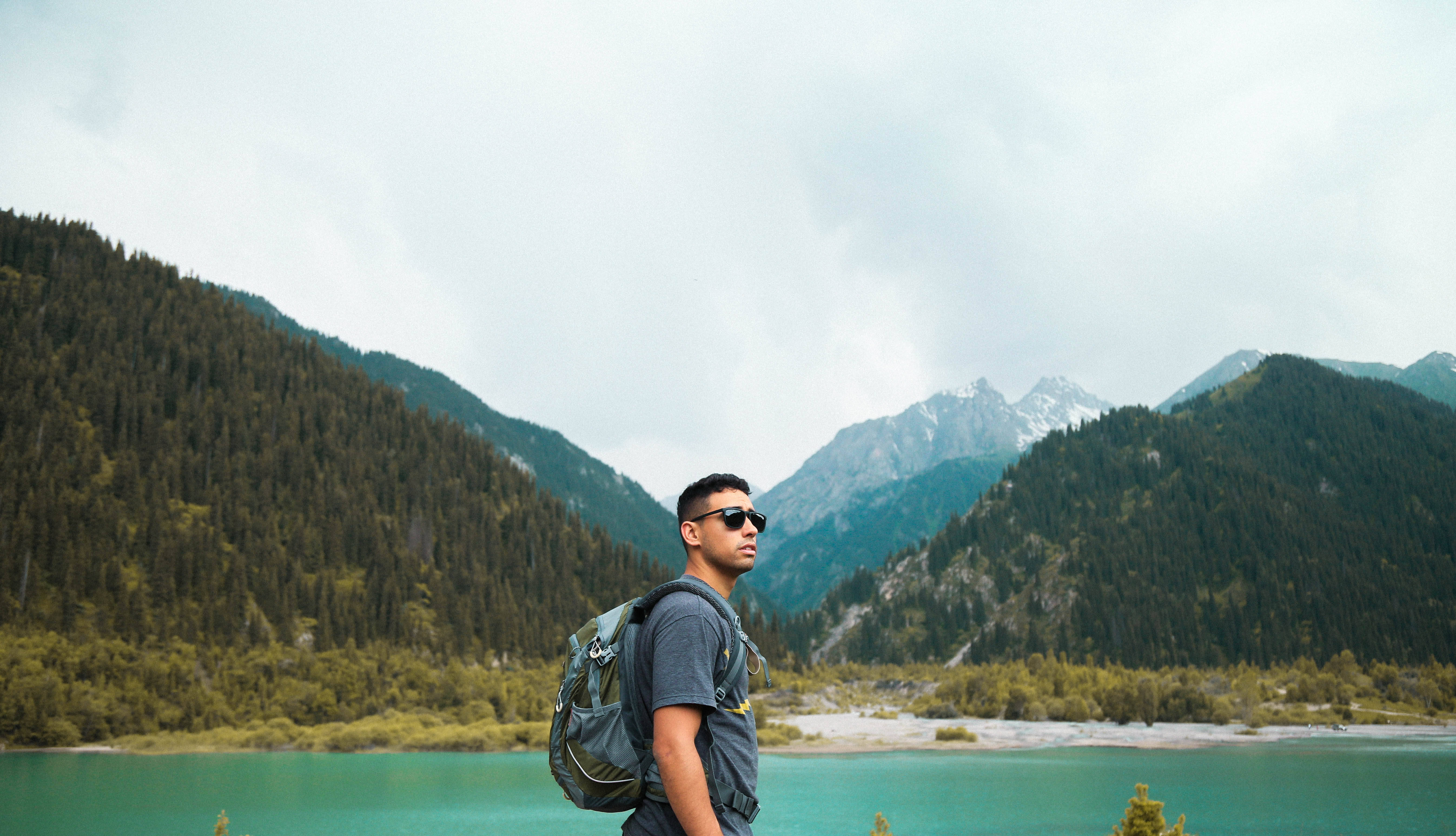 Kazakhstan 桌布 壁紙 — 一個背著背包、戴著太陽眼鏡的男人站在一個碧綠色的湖邊，周圍是森林覆蓋的山脈，天空多雲。