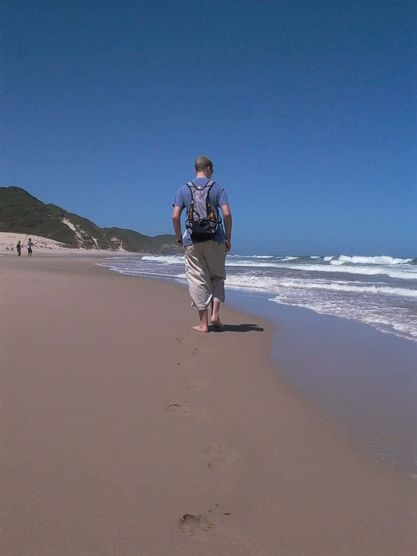 Beach Walk 桌布 壁紙 — 一個背著背包的男人赤腳走在寬闊的沙灘上，天空清澈湛藍，海浪翻滾而來。
