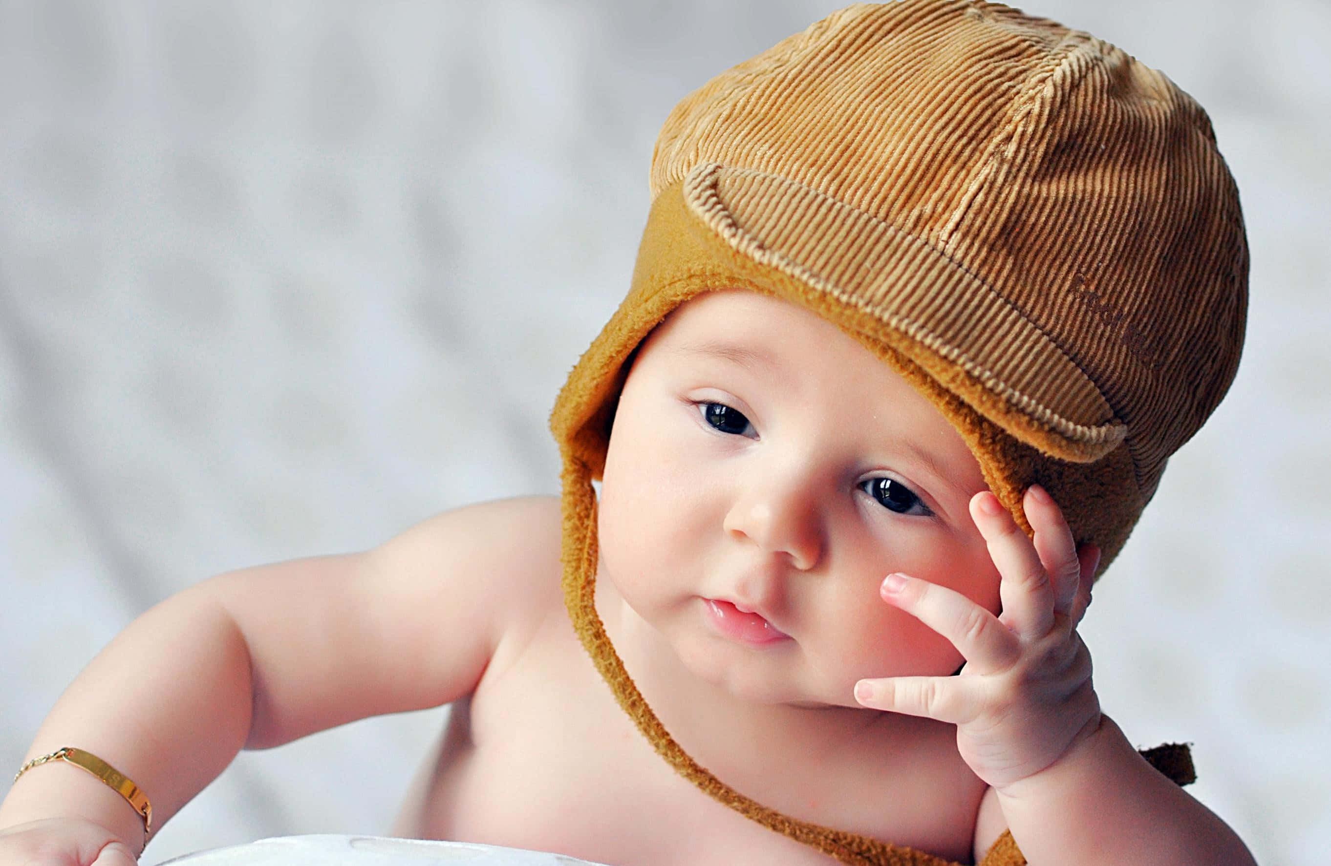 Baby Boy 桌布 壁紙 — 一個胖嘟嘟的嬰兒戴著芥末黃燈芯絨帽子，好奇地凝視著，同時將手放在臉頰附近。