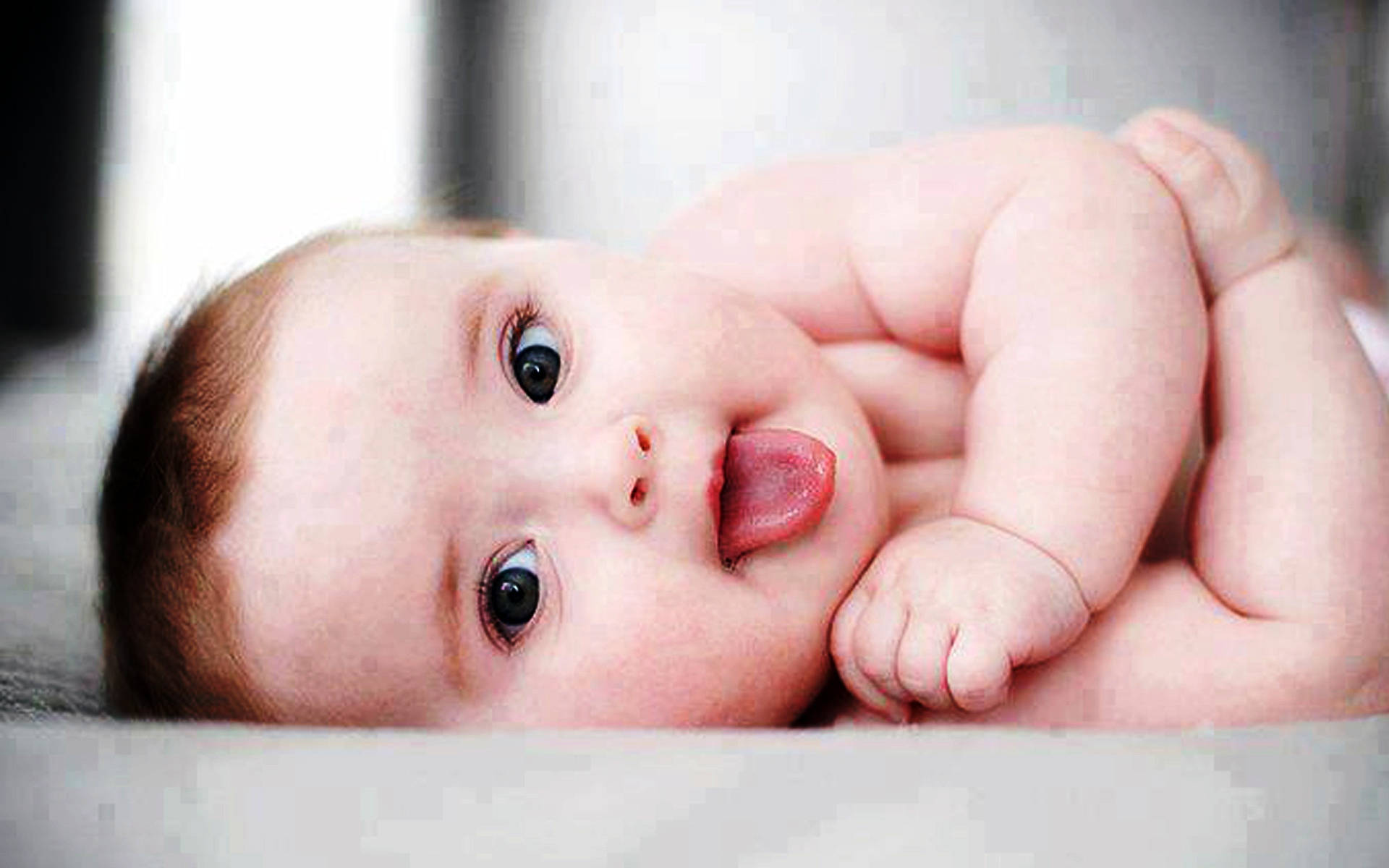 Funny Baby 桌布 壁紙 — 一個胖嘟嘟的寶寶，有著一雙大黑眼睛，在柔軟的表面上仰躺著伸出舌頭。