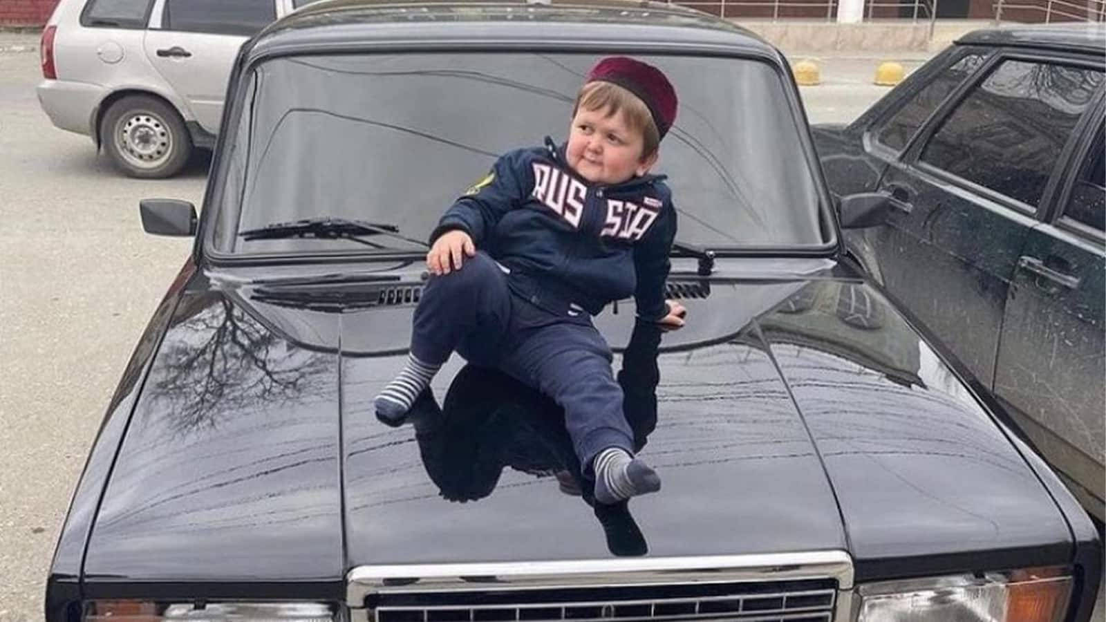 Celebrity 桌布 壁紙 — 一個胖嘟嘟的幼兒，穿著海軍藍色「RUS 517」連帽衫和紅色帽子，自信地坐在一輛閃亮黑色汽車的引擎蓋上。