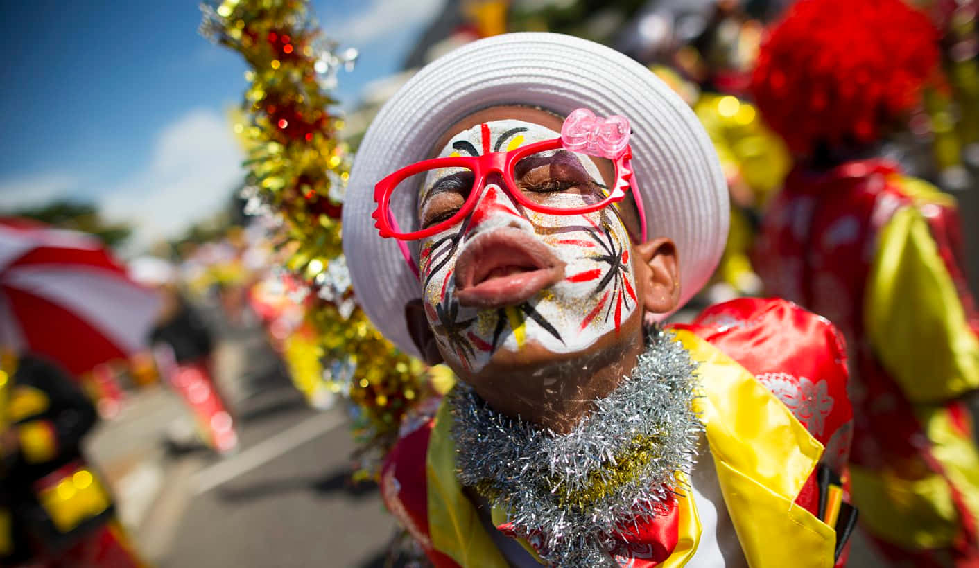 African Culture 桌布 壁紙 — 一個臉上塗滿鮮豔顏料、戴著紅色眼鏡的小孩，在明媚陽光下的節日街頭遊行中歡樂地綻放笑容。