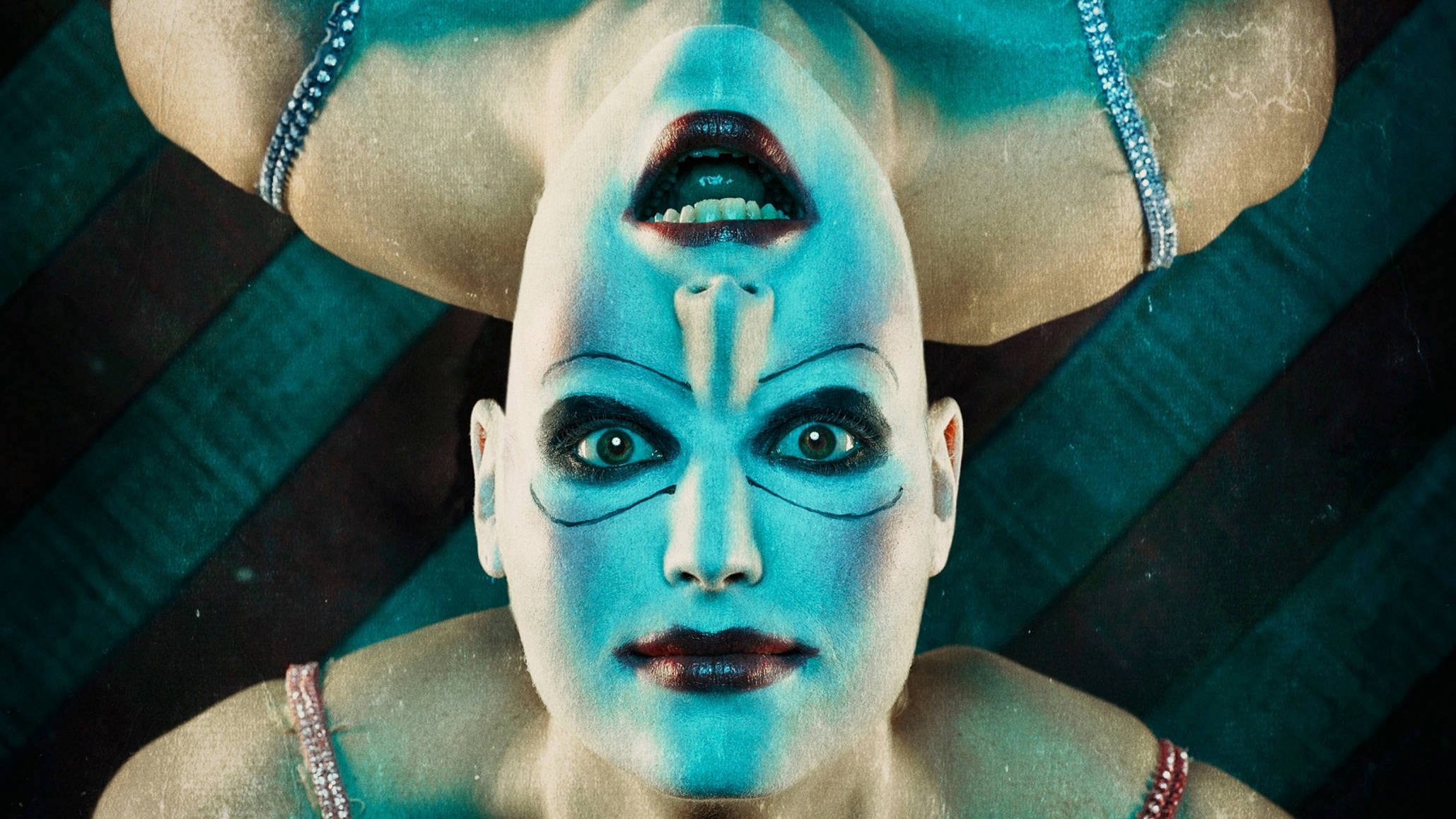 American Horror Story 桌布 壁紙 — 一個臉上塗著藍色顏料、畫著深色眼妝的人，戴著鑲有寶石的帶子，在青綠色條紋背景上向上凝視。