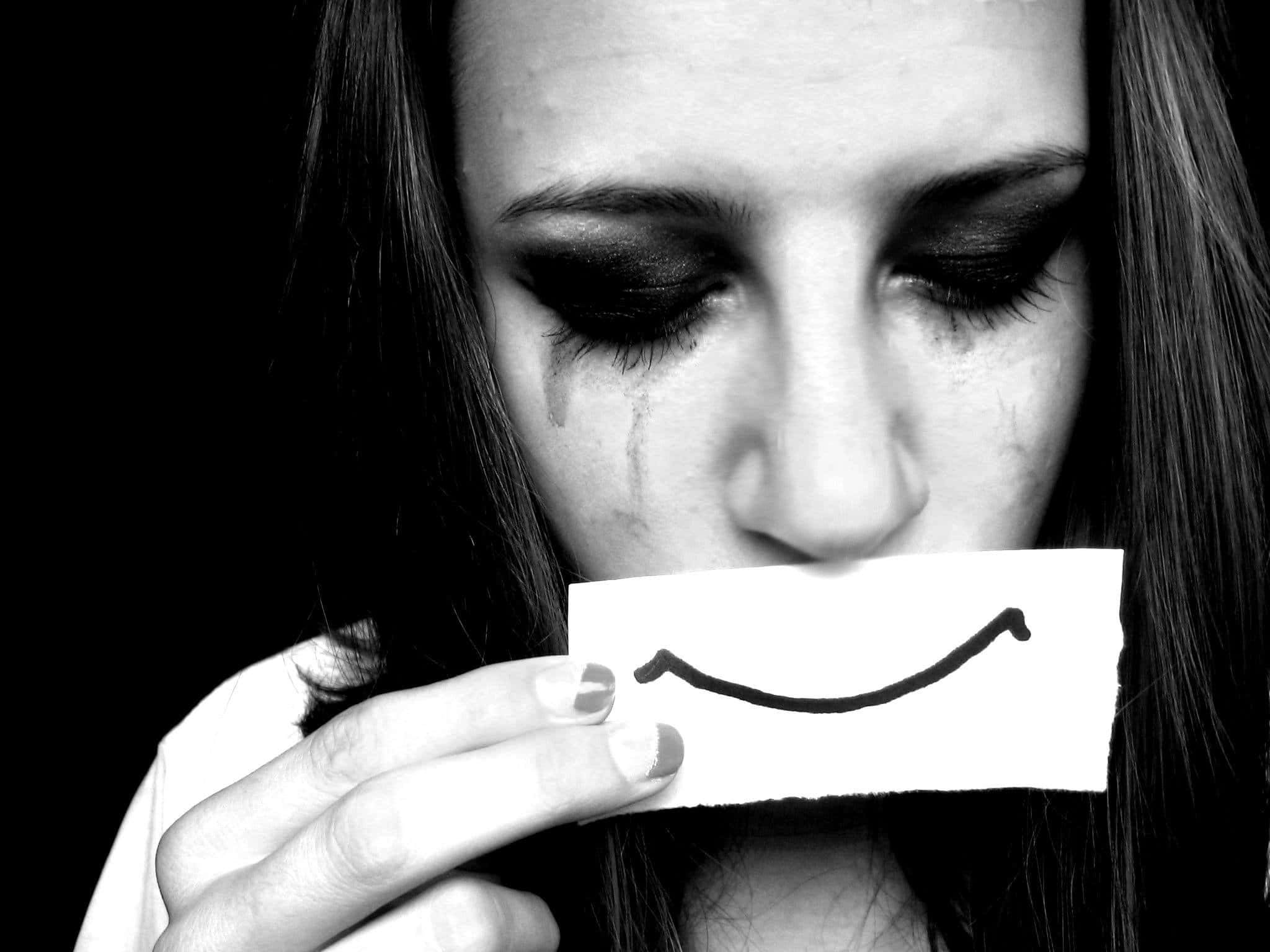 Emotional 桌布 壁紙 — 一個臉上帶有淚痕的女人，在黑色背景前將一個紙製笑臉放在嘴巴上。