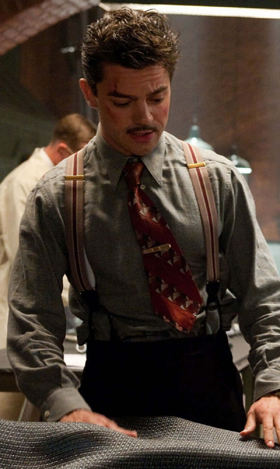 Howard Stark 桌布 壁紙 — 一個臉部瘀傷、留鬍子的男人，穿著吊帶褲和圖案領帶，在光線昏暗的房間裡俯身於一個有紋理的表面上。