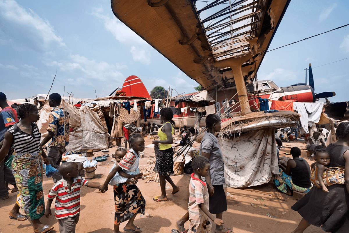 Africa 桌布 壁紙 — 一個臨時定居點的熱鬧日間場景，孩子們在破舊的庇護所附近玩耍，頭頂是明亮的藍天。