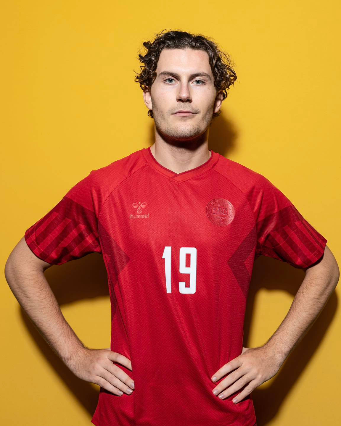 Football Players 桌布 壁紙 — 一個自信的年輕男子身穿紅色足球球衣（19號），雙手叉腰，站在鮮黃色背景前。