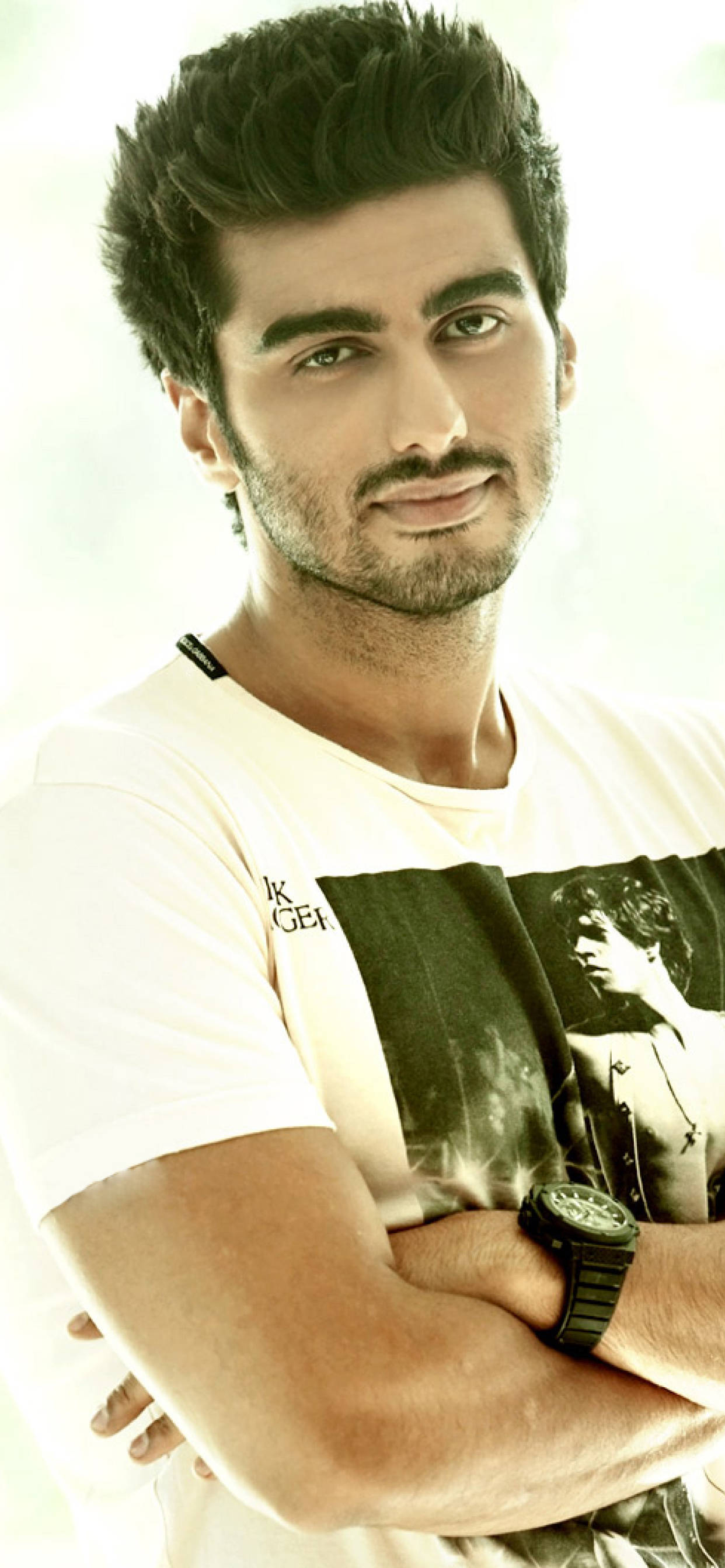Arjun Kapoor 桌布 壁紙 — 一個自信的男人，留著時尚的深色頭髮和鬍鬚，穿著圖案T恤和黑色手錶，雙臂交叉站在明亮的背景前。