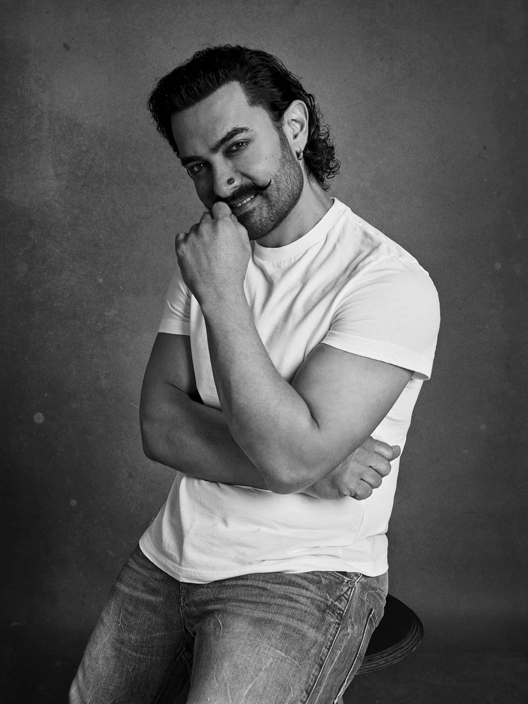 Aamir Khan 桌布 壁紙 — 一個自信的男人，留著鬍子並戴著耳環，坐在凳子上，手托著下巴，在一張充滿氛圍的黑白攝影棚肖像照中。