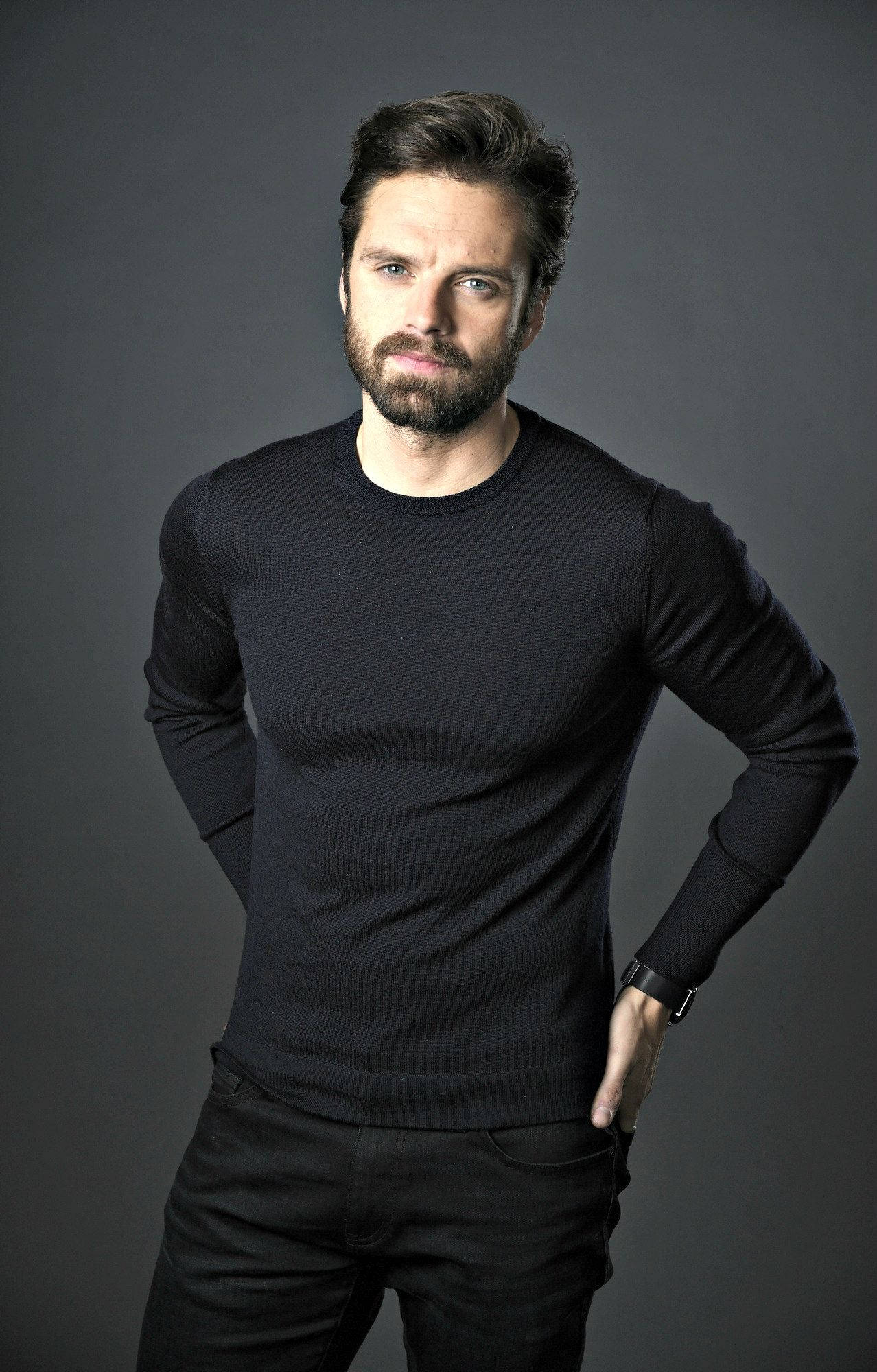 Sebastian 桌布 壁紙 — 一個自信的男人，留著鬍鬚和深色頭髮，站在灰色背景前，穿著黑色長袖襯衫和深色褲子。