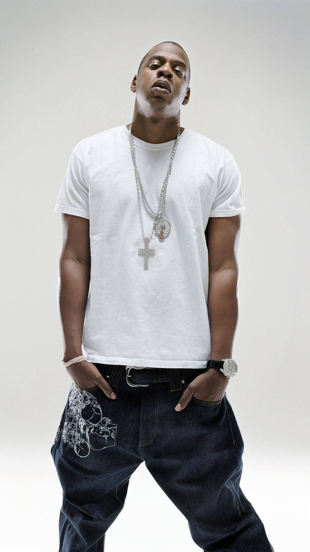 Jay-z 桌布 壁紙 — 一個自信的男人，穿著白色T恤和深色牛仔褲，雙手插口袋，站在純色攝影棚背景前擺姿勢。