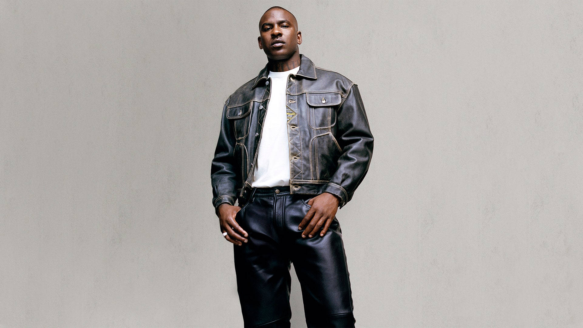 Skepta 桌布 壁紙 — 一個自信的男人，穿著黑色皮夾克和褲子，雙手插在口袋裡，站在純灰色的攝影棚牆邊。