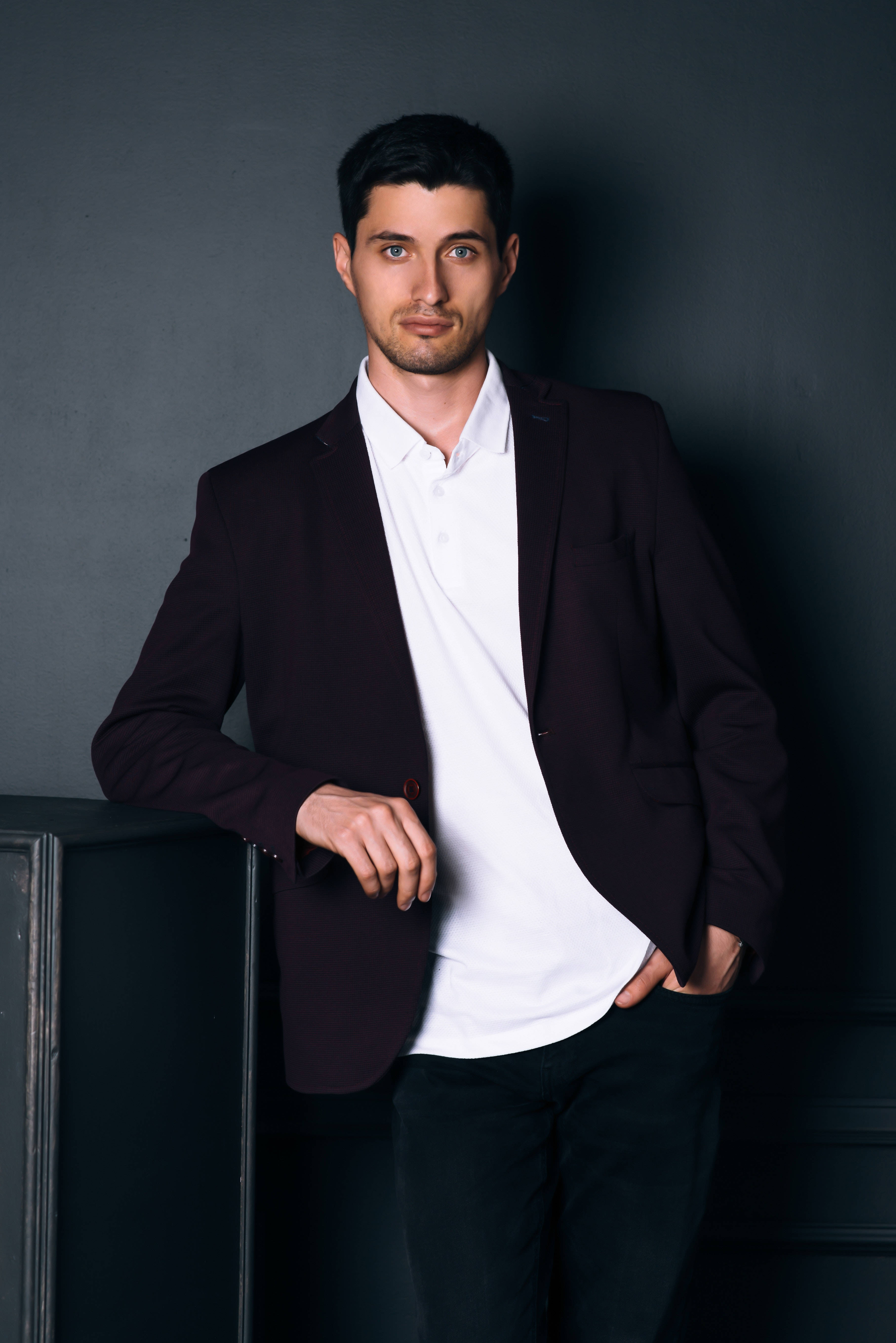 male model 桌布 壁紙 — 一個自信的男人，身穿栗色西裝外套和白色襯衫，靠著灰色牆前的深色櫥櫃。