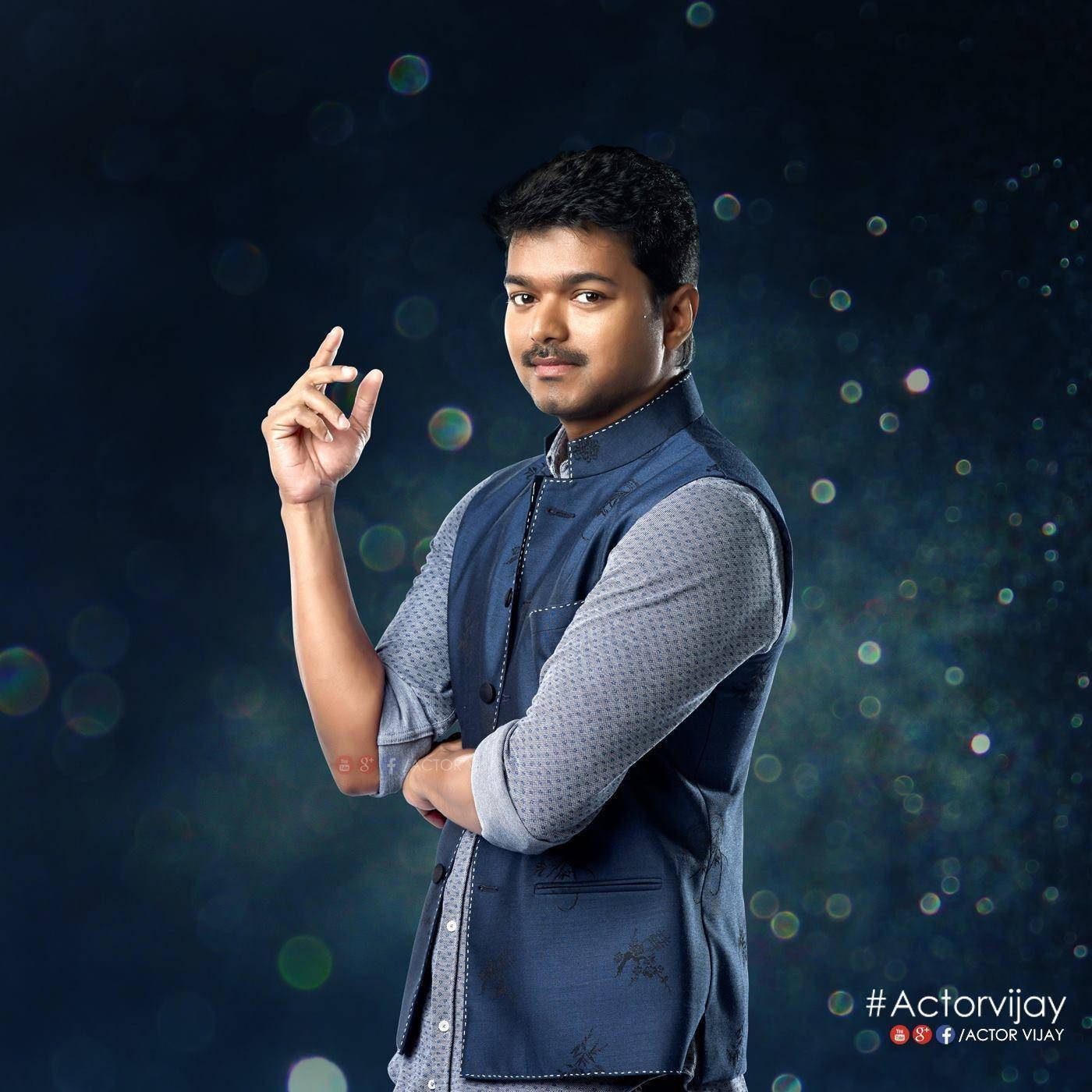vijay HD 桌布 壁紙 — 一個自信的男人，身穿藍色背心和圖案襯衫，在一個有發光球體的暗色散景背景前擺姿勢。