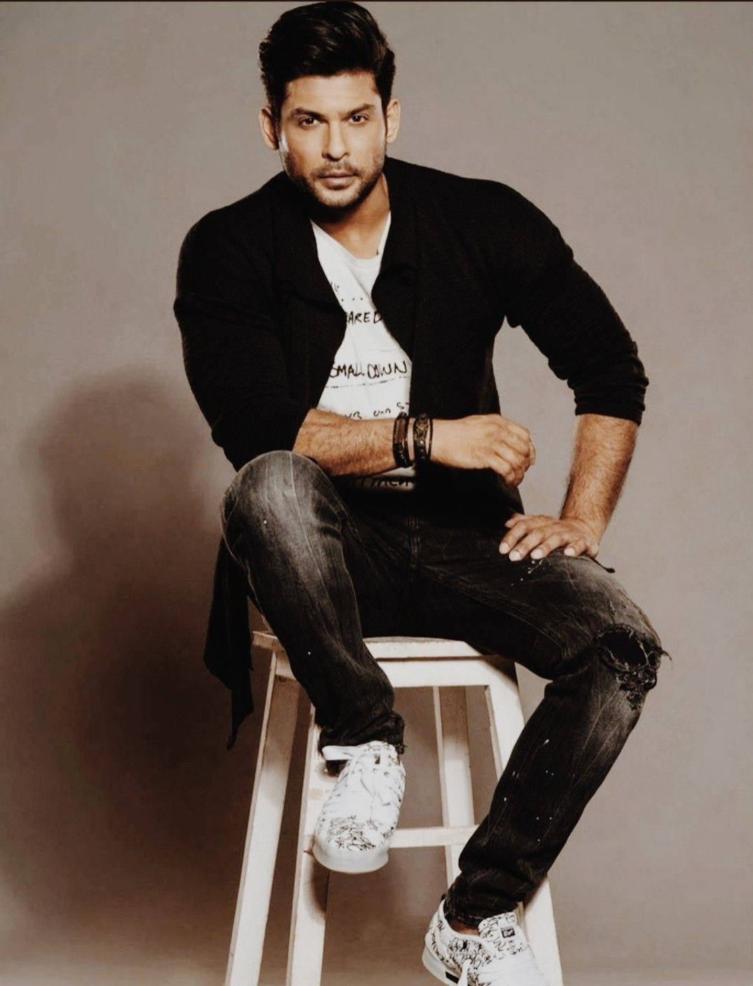 Sidharth Shukla 桌布 壁紙 — 一個自信的男人，頭髮造型整齊，隨意地坐在白色凳子上，穿著黑色開襟毛衣、破洞牛仔褲和白色運動鞋，背景是簡單的攝影棚。