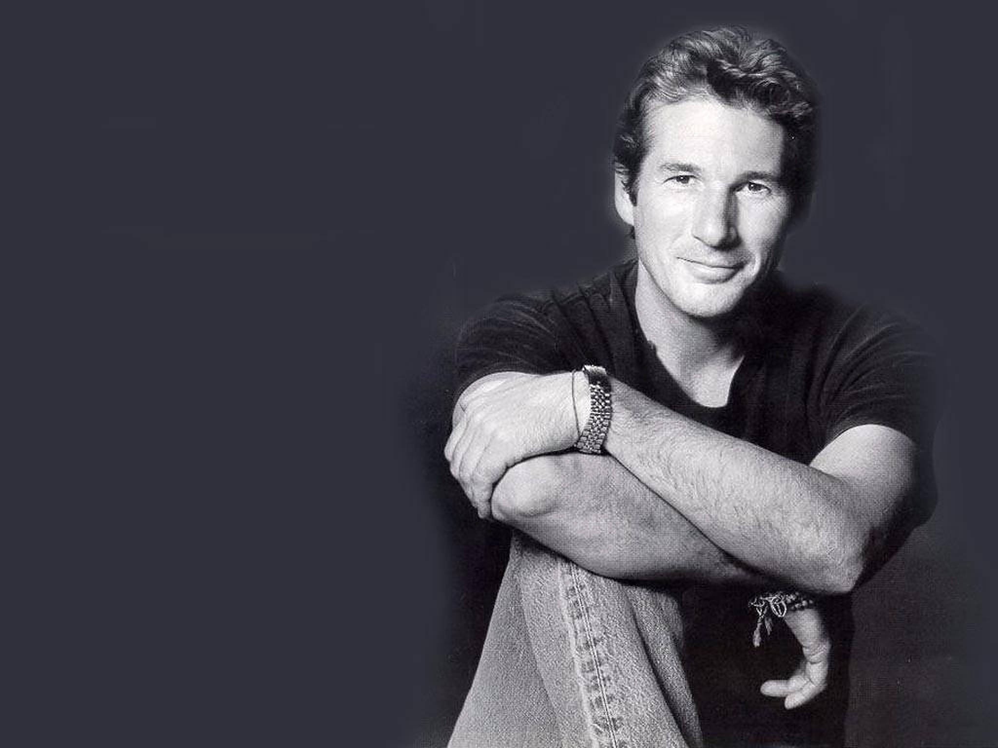 Richard Gere 桌布 壁紙 — 一個自信的男人交叉雙臂，在黑暗的工作室背景下溫暖地微笑，經典黑白肖像。