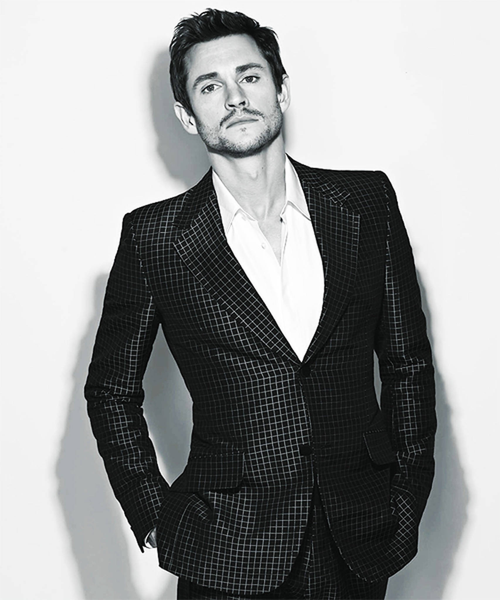 Hugh Dancy 桌布 壁紙 — 一個自信的男人穿著格紋西裝，站在樸素的牆邊，雙手插在口袋裡，呈現出一張戲劇性的黑白肖像照。