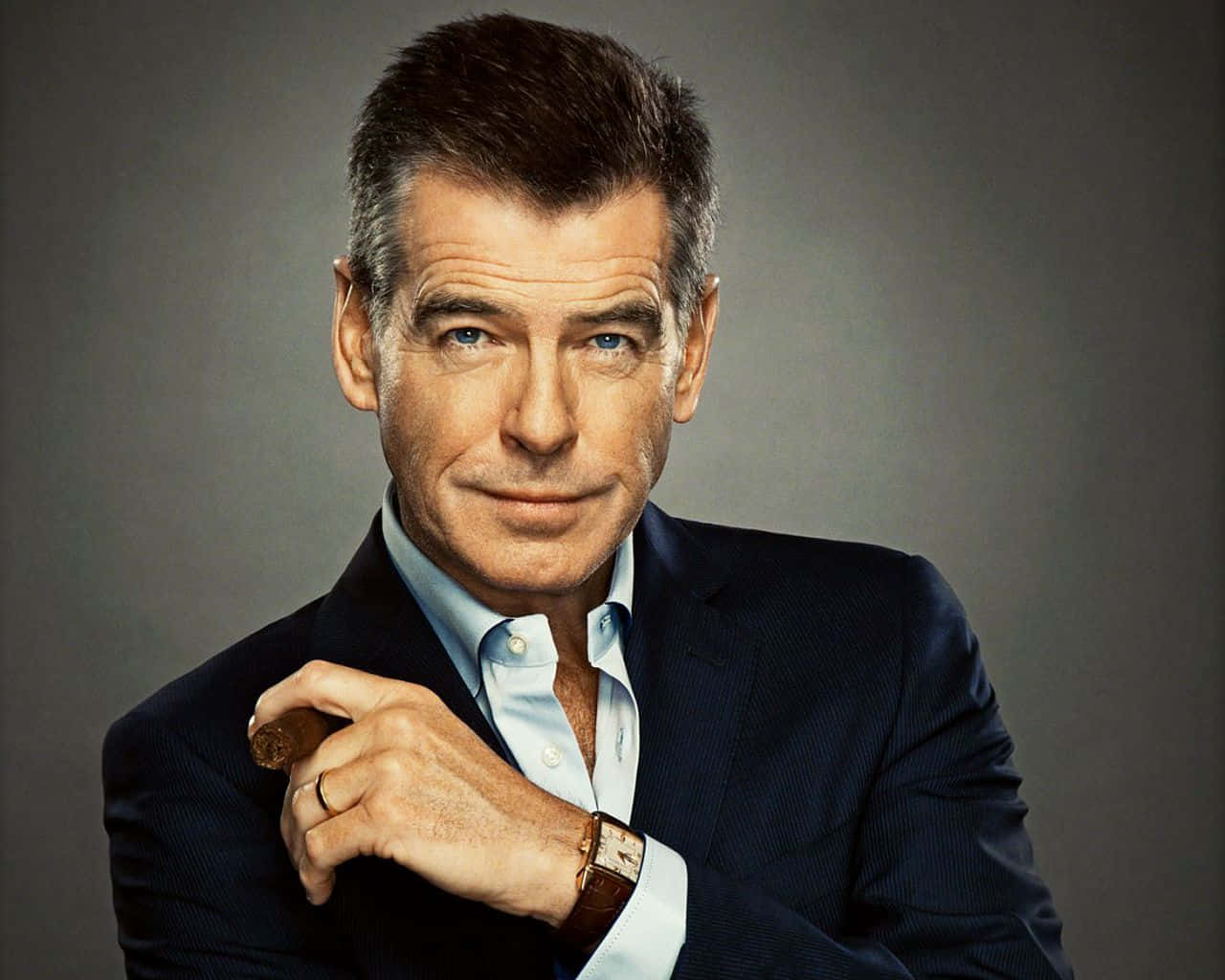 pierce brosnan 桌布 壁紙 — 一個自信的男人穿著深色西裝，在柔和的灰色背景前拿著雪茄，有著強烈的藍眼睛。