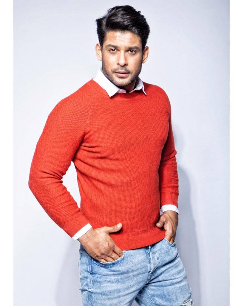 Sidharth Shukla 桌布 壁紙 — 一個自信的男人穿著紅色毛衣和藍色牛仔褲，雙手插袋，在純灰色背景前擺姿勢。