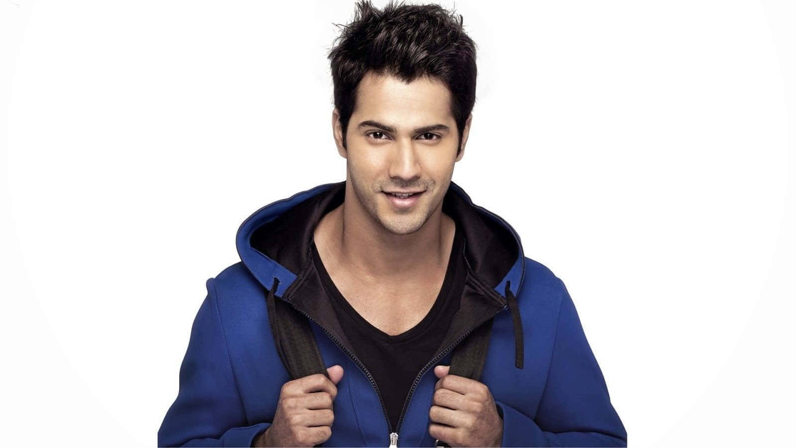 Varun Dhawan 桌布 壁紙 — 一個自信的男人穿著藍色連帽衫，在純白背景上拉開拉鍊微笑。