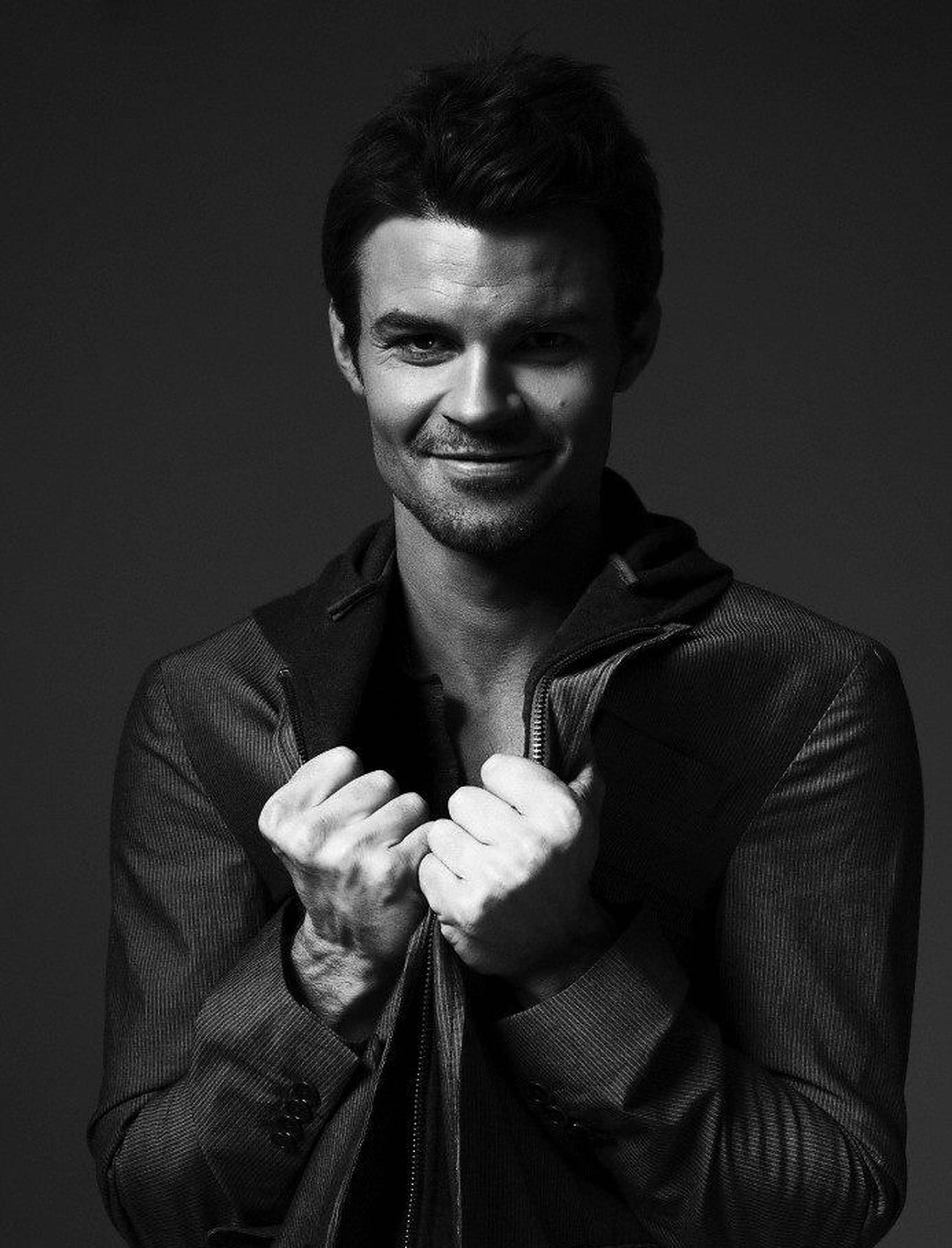 Daniel Gillies 桌布 壁紙 — 一個自信的男人穿著質感外套，在黑暗的攝影棚背景下微笑著抓住拉鍊。